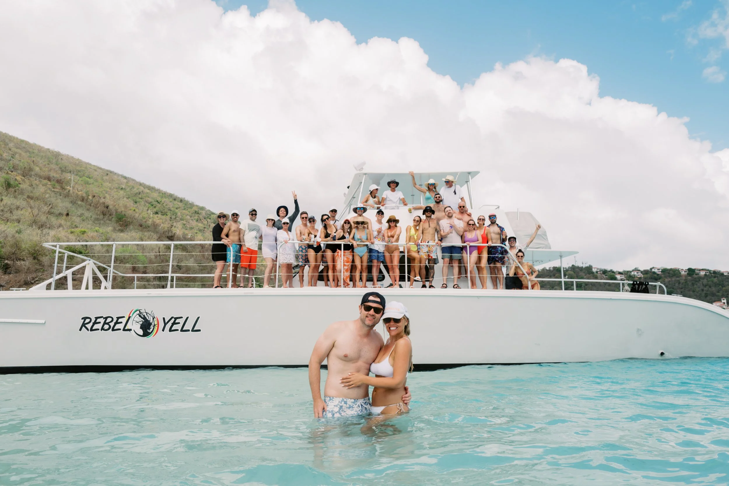 A group of people on a yacht named 'REBEL YELL' near a shoreline, with two people in swimsuit in the water in the foreground, and a hilly landscape under a partly cloudy sky in the background.