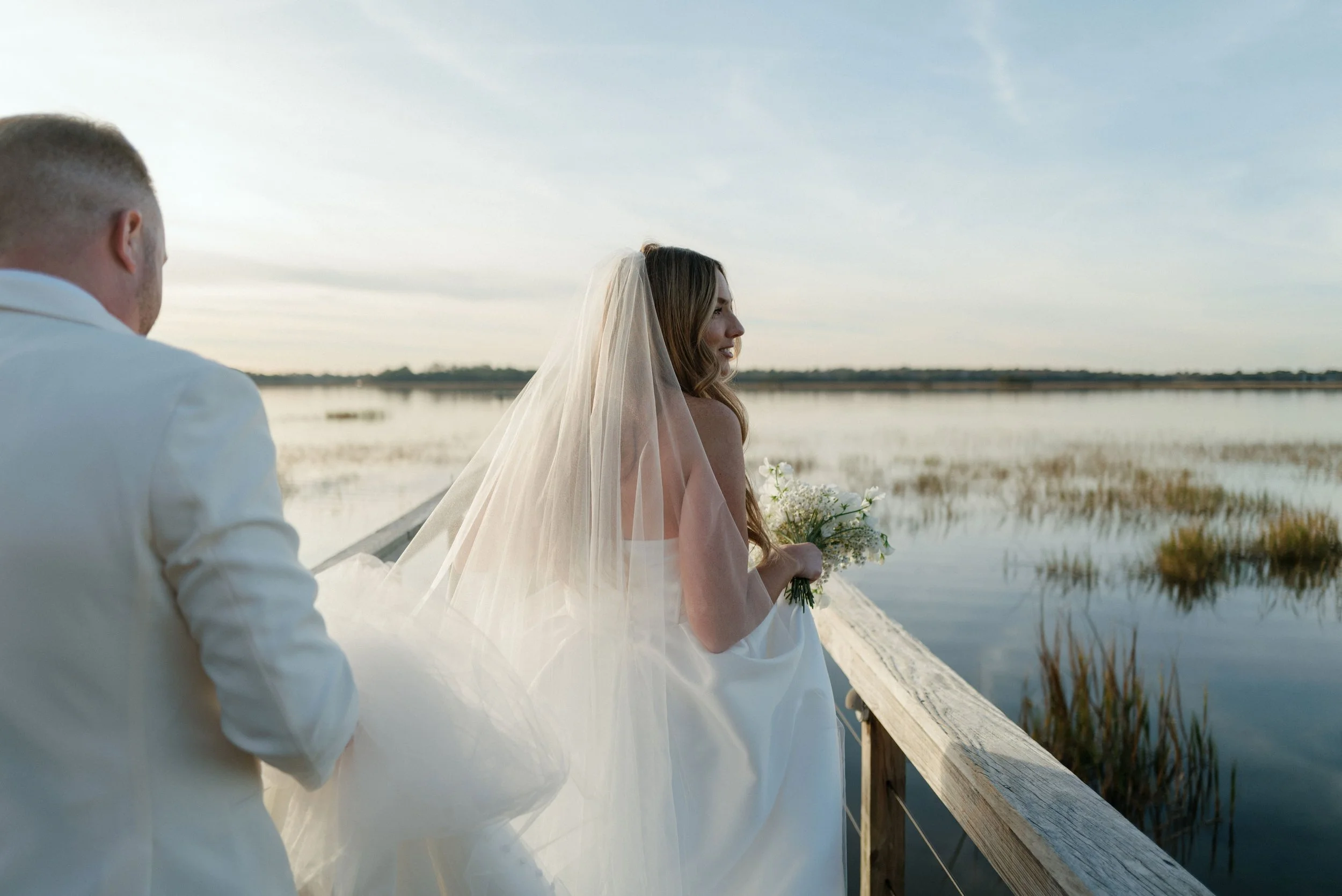 A bride holding a bouquet of white flowers and smiling, standing on a wooden dock by a calm body of water, with a groom nearby, during a sunny day