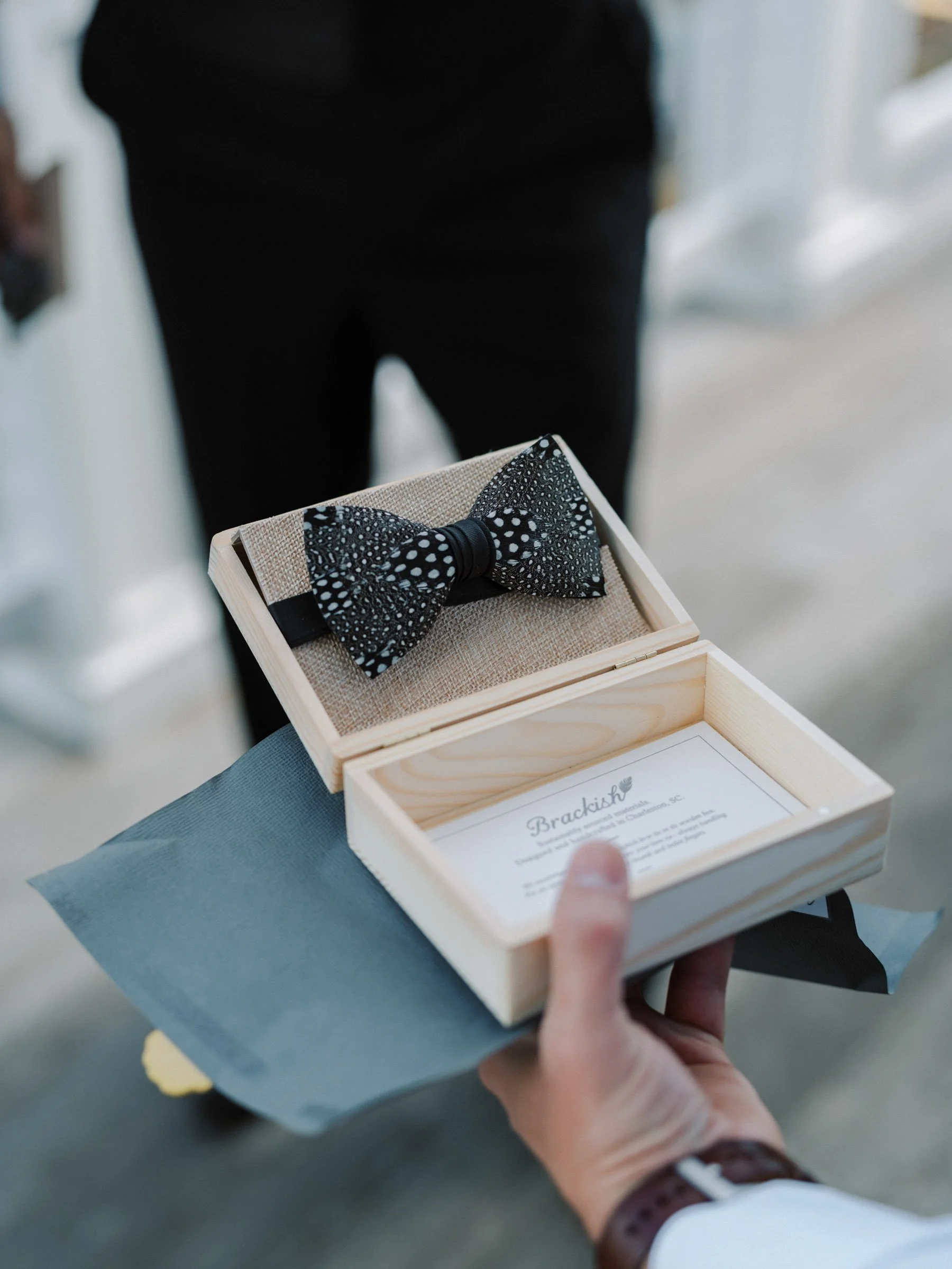 Person holding an open wooden box with a black patterned bow tie and a card inside.
