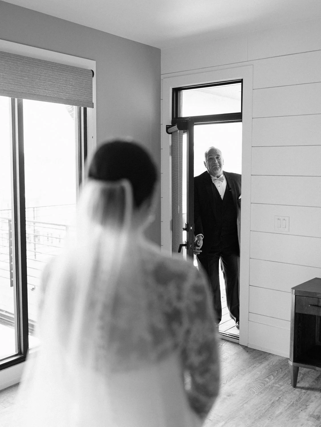 A bride in a wedding dress with a tattoo on her shoulder, standing inside a room, looks at an older man in a tuxedo standing on a balcony outside the door.