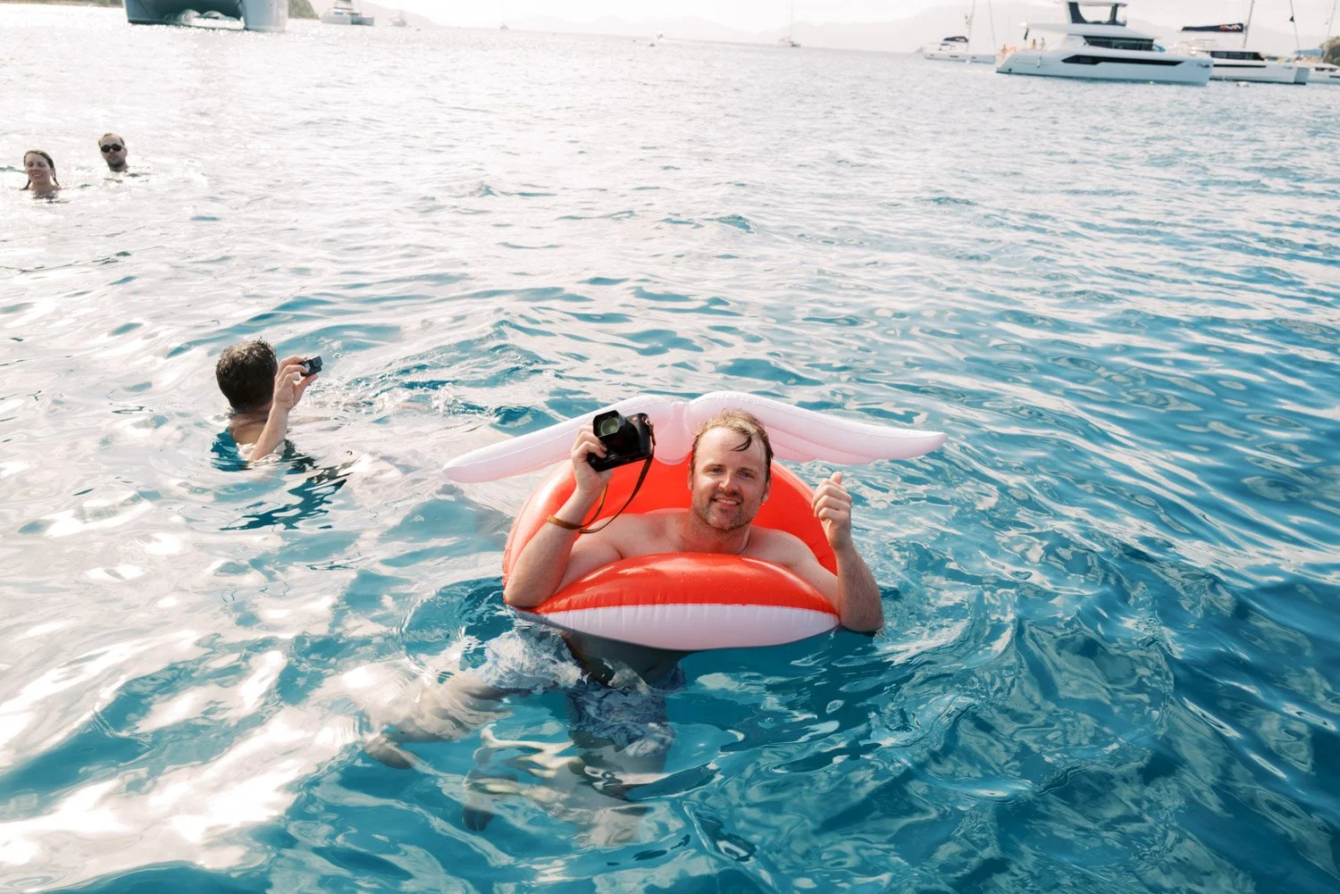 Man floating in water on an inflatable with a unicorn horn, holding a camera, with other people swimming nearby and yachts in the background.