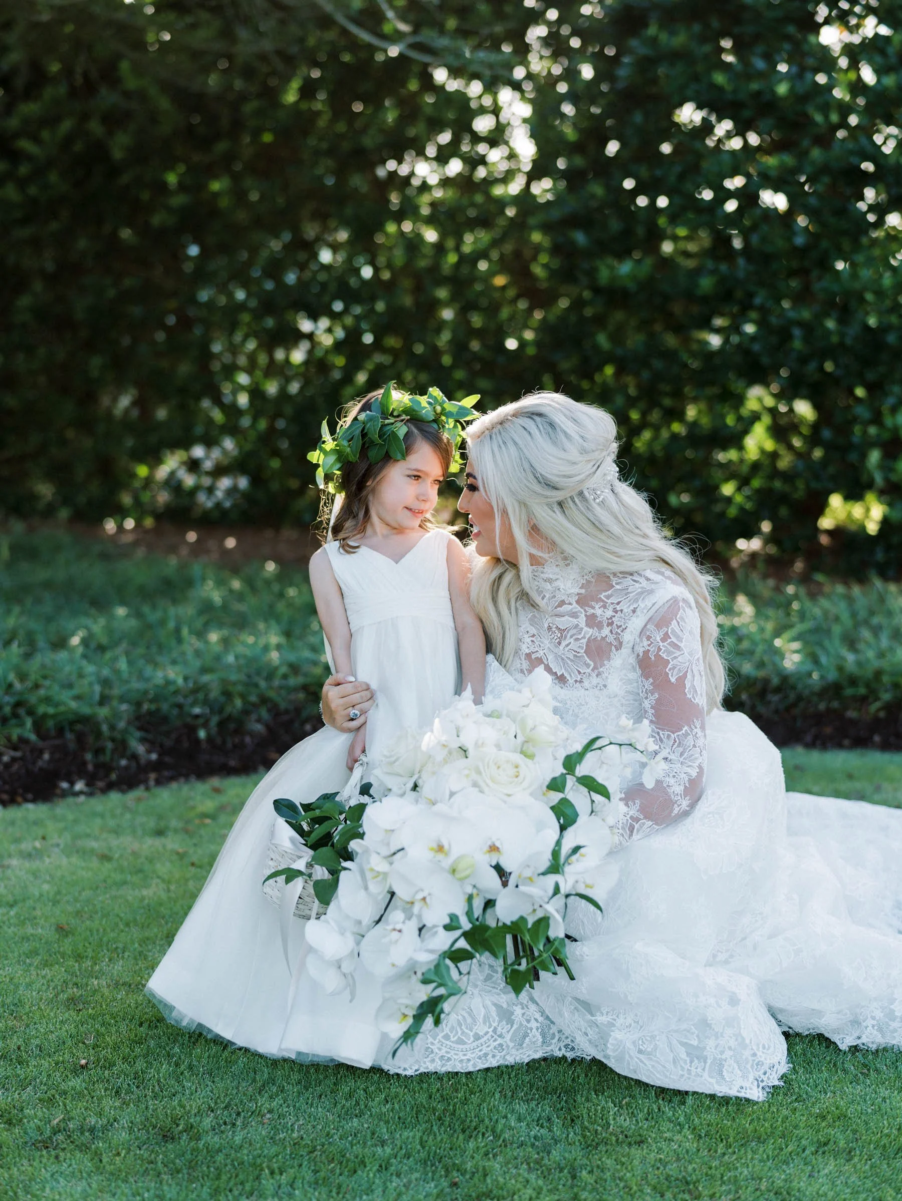 A woman in a lace wedding dress and a young girl with a floral crown are kneeling together on a grassy area, smiling at each other, with a bouquet of white flowers in front of them, outdoors with green trees in the background.