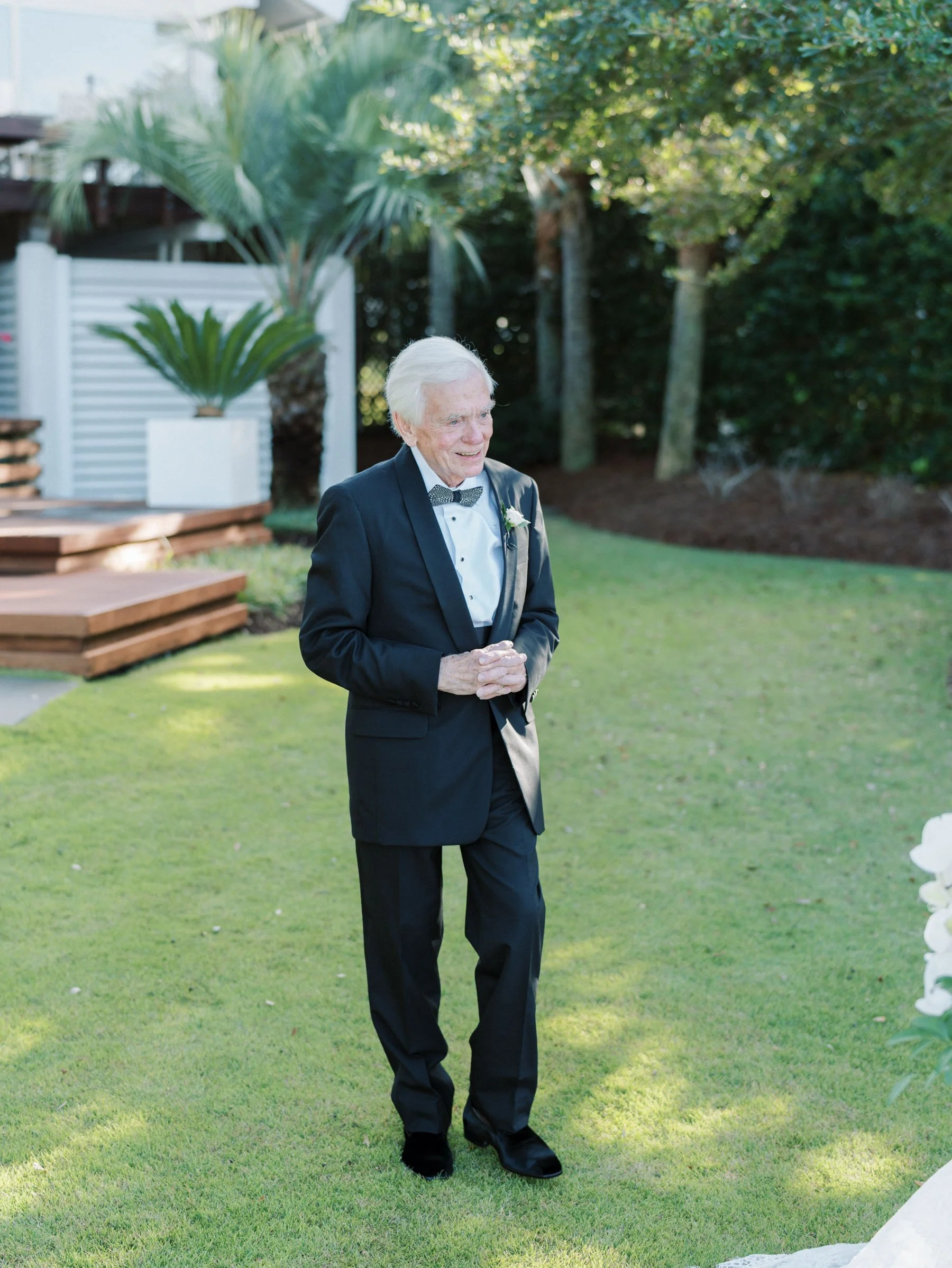 An elderly man in a tuxedo with a bow tie and a white flower boutonniere standing on a lawn during a garden wedding ceremony.
