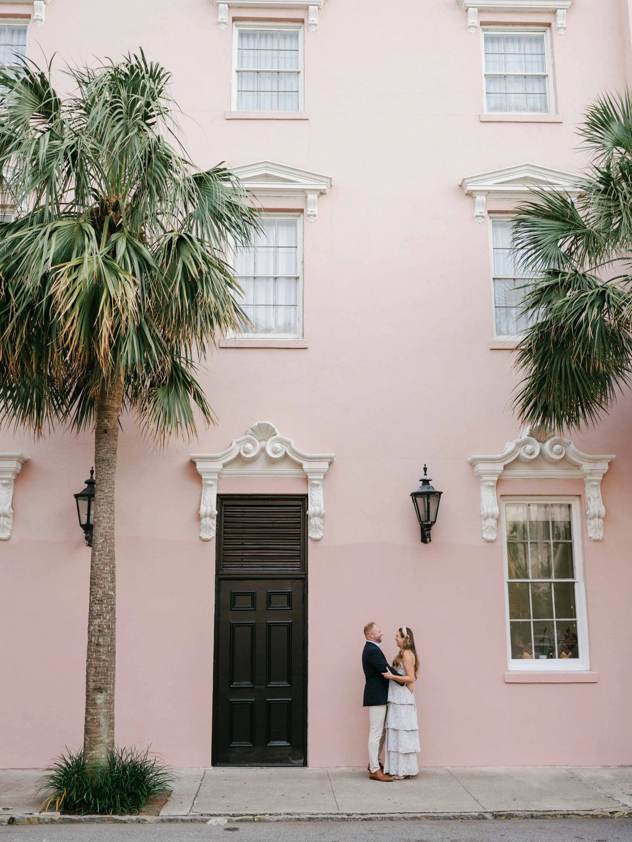 A couple stands together on a sidewalk in front of a pink building with tall palm trees, white window frames, decorative trim, and black lanterns.
