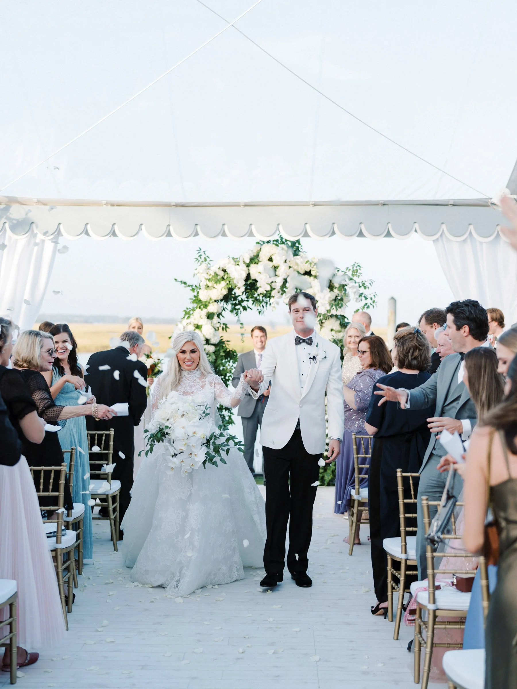 Bride and groom walking down the aisle at an outdoor wedding ceremony under a tent, surrounded by guests.