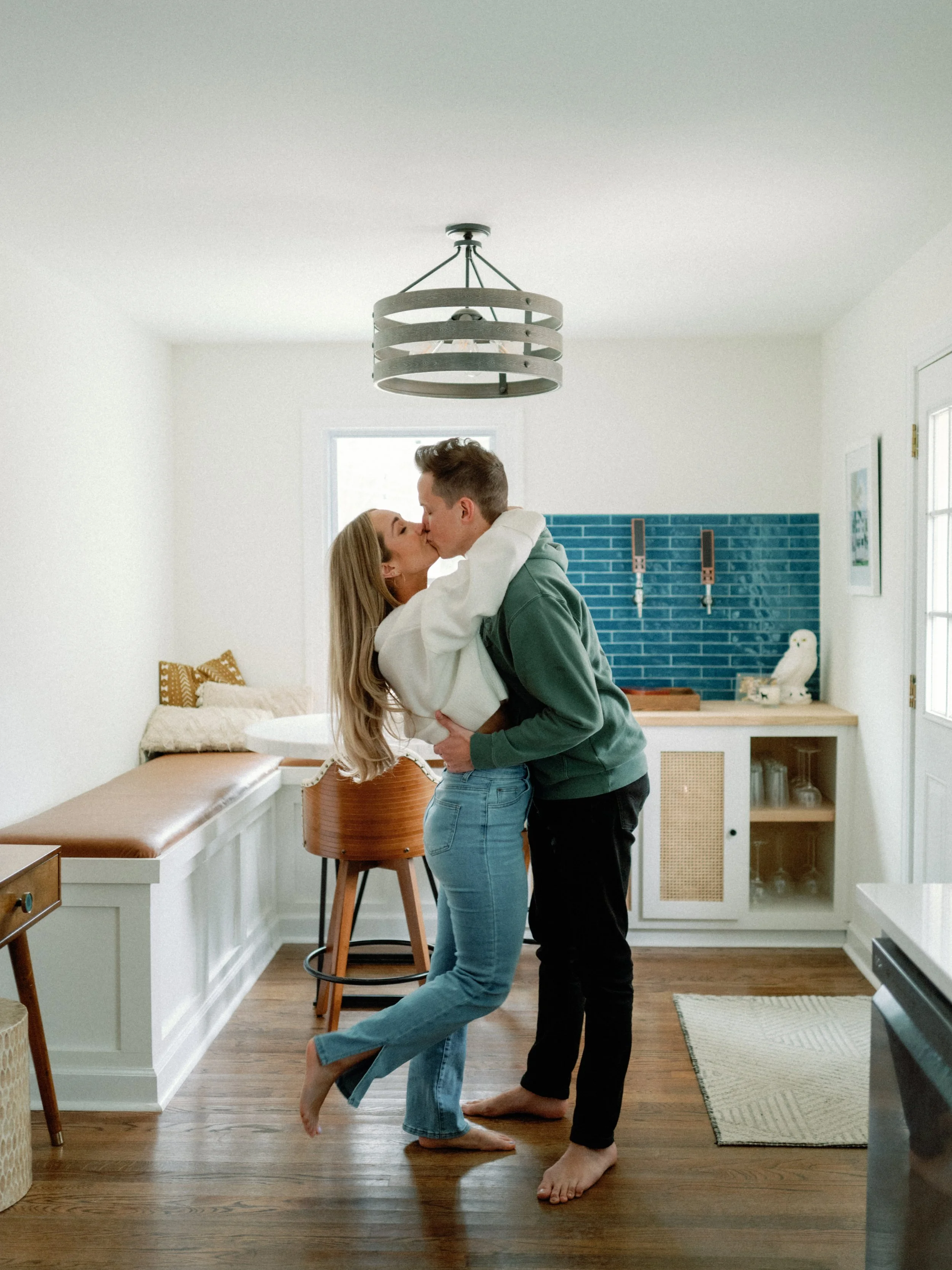 A couple embracing and kissing in a kitchen with wooden flooring, white walls, a blue tiled backsplash, and a window.