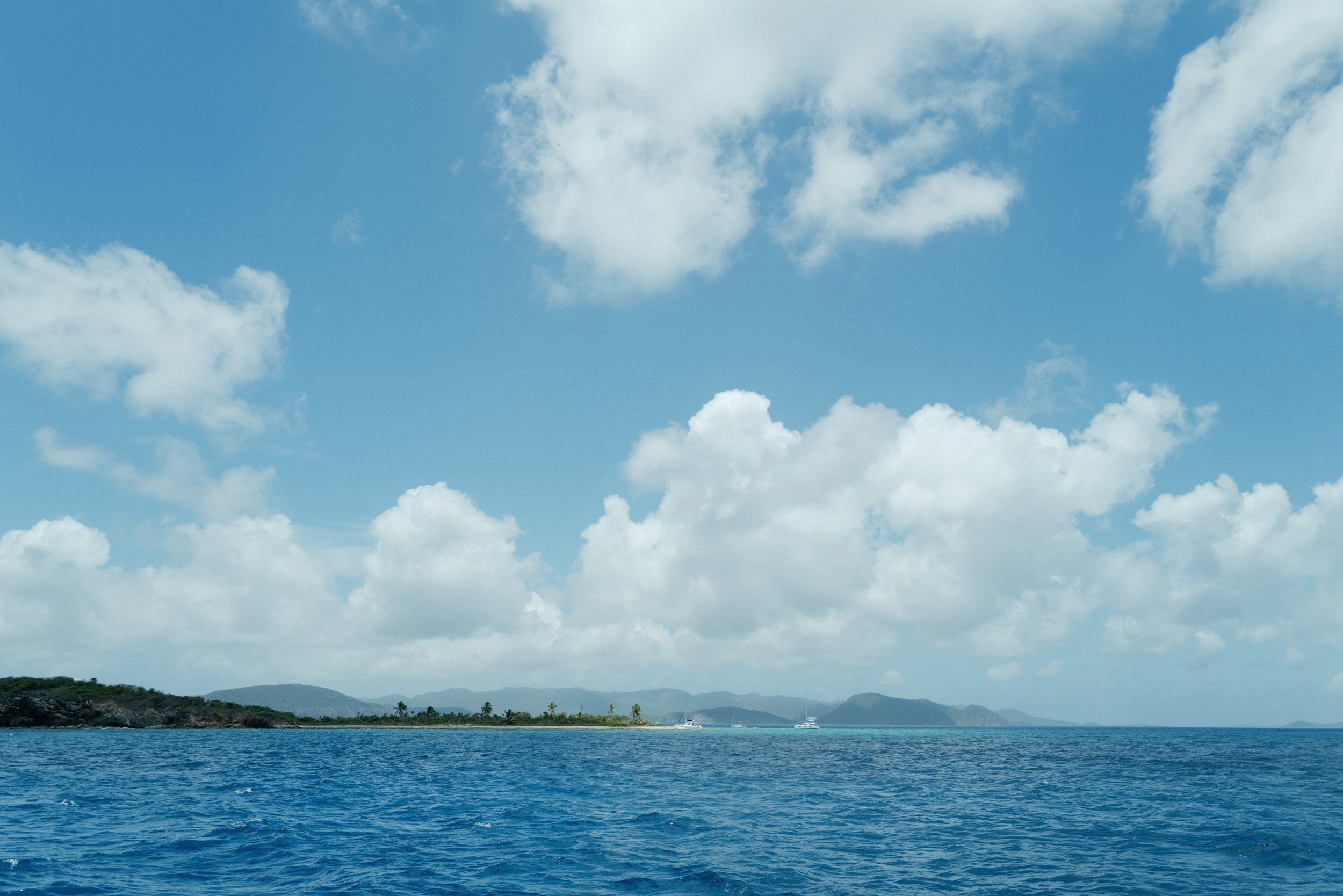 A scenic view of the British Virgin Island ocean with blue waters, a distant shoreline with trees, and a partly cloudy sky.