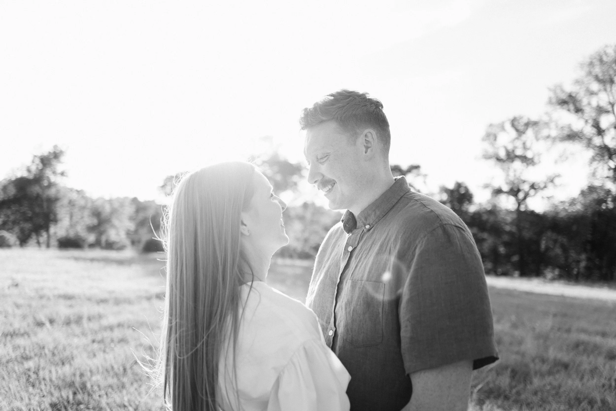 A black and white photo of a couple smiling and looking into each other's eyes outdoors on a sunny day, with trees in the background.