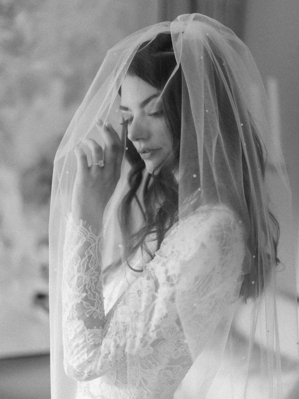 A woman with long dark hair in a wedding dress, covering her face with a sheer veil, looking down thoughtfully.