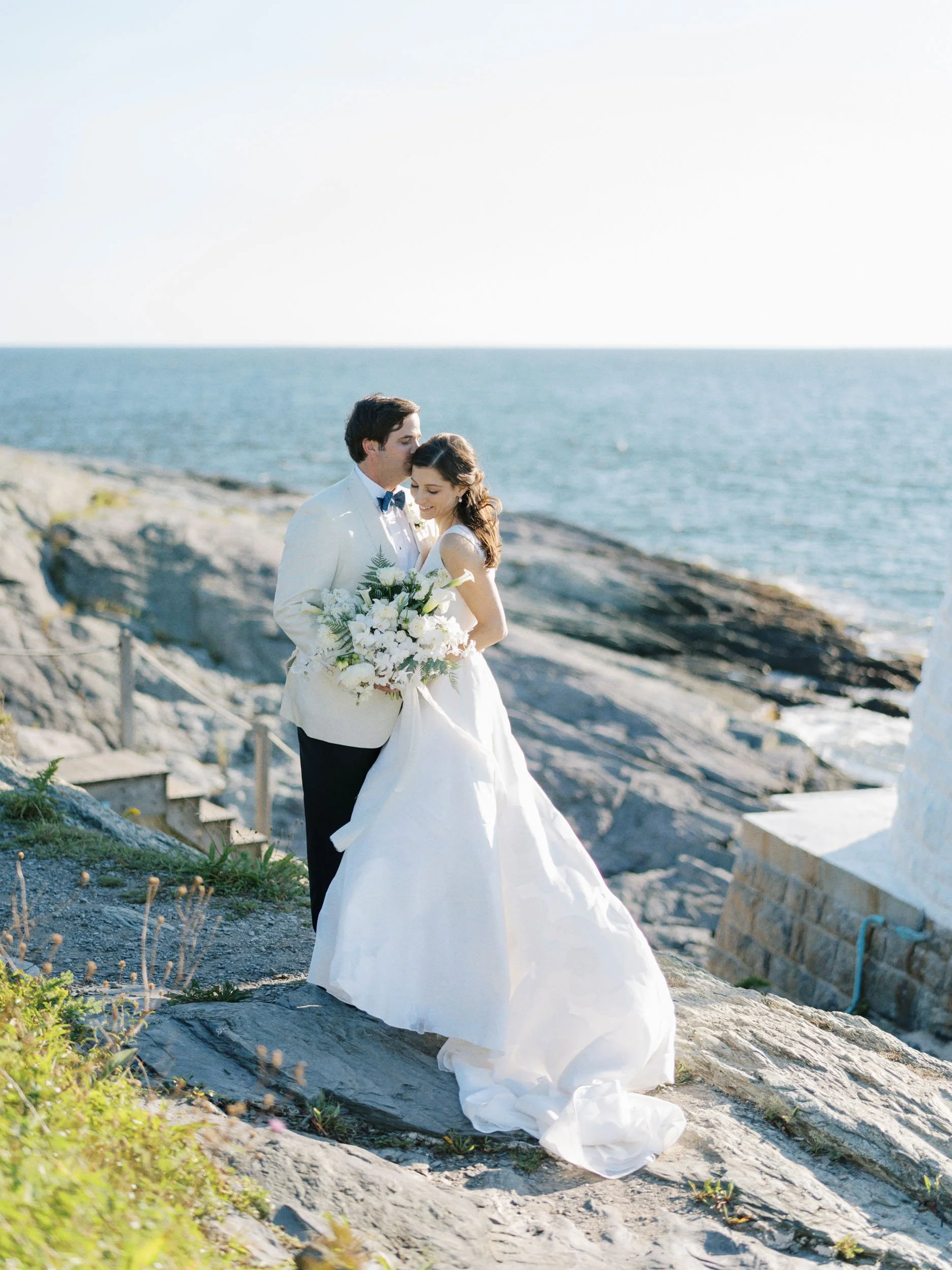 A bride and groom stand on rocky terrain near the ocean, embracing during their wedding, with the bride holding a bouquet of white flowers.