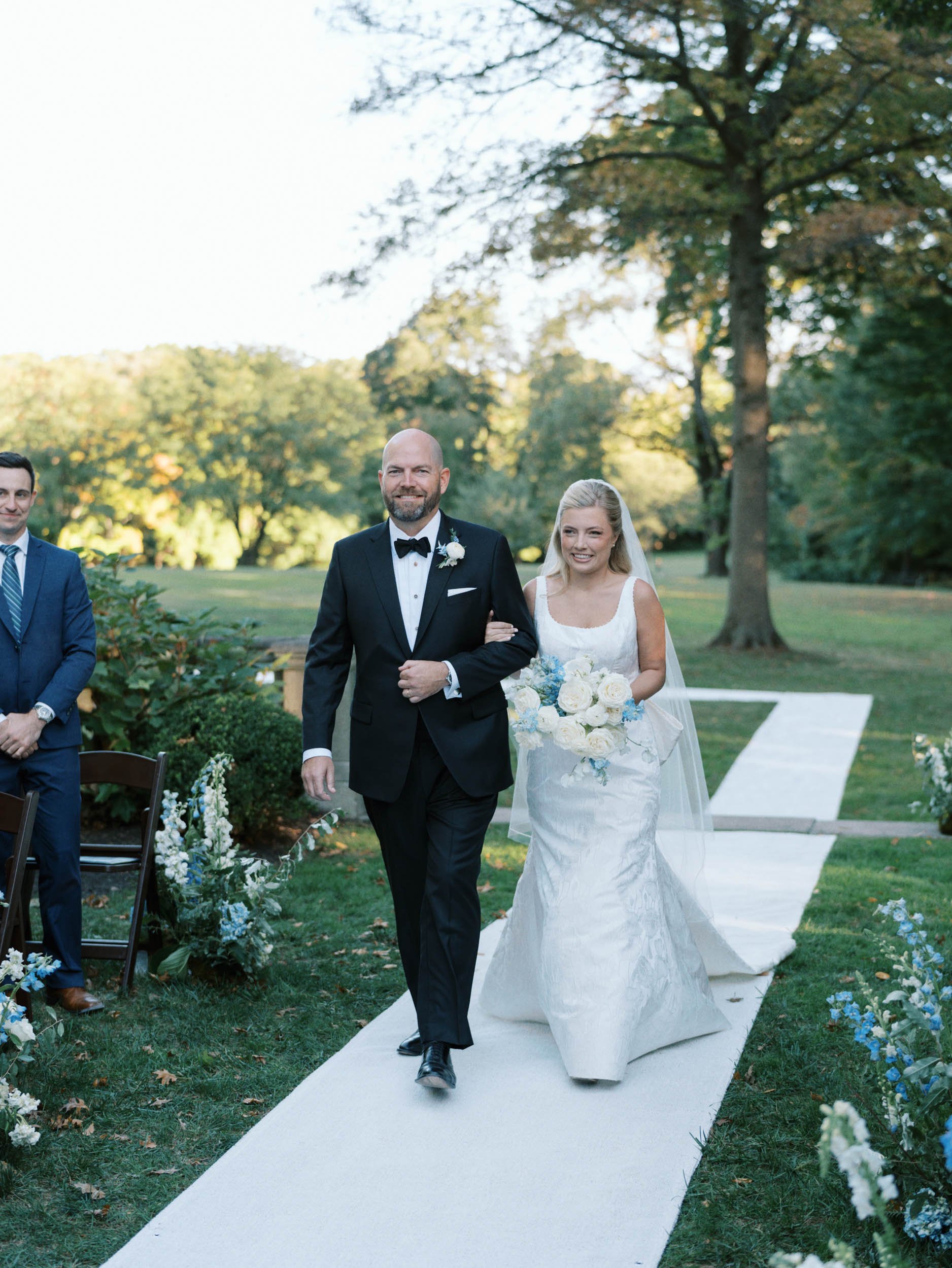 A bride walking down the aisle with her father at an outdoor wedding ceremony. The bride is wearing a white gown and holding a bouquet. The background shows trees and a grassy area.
