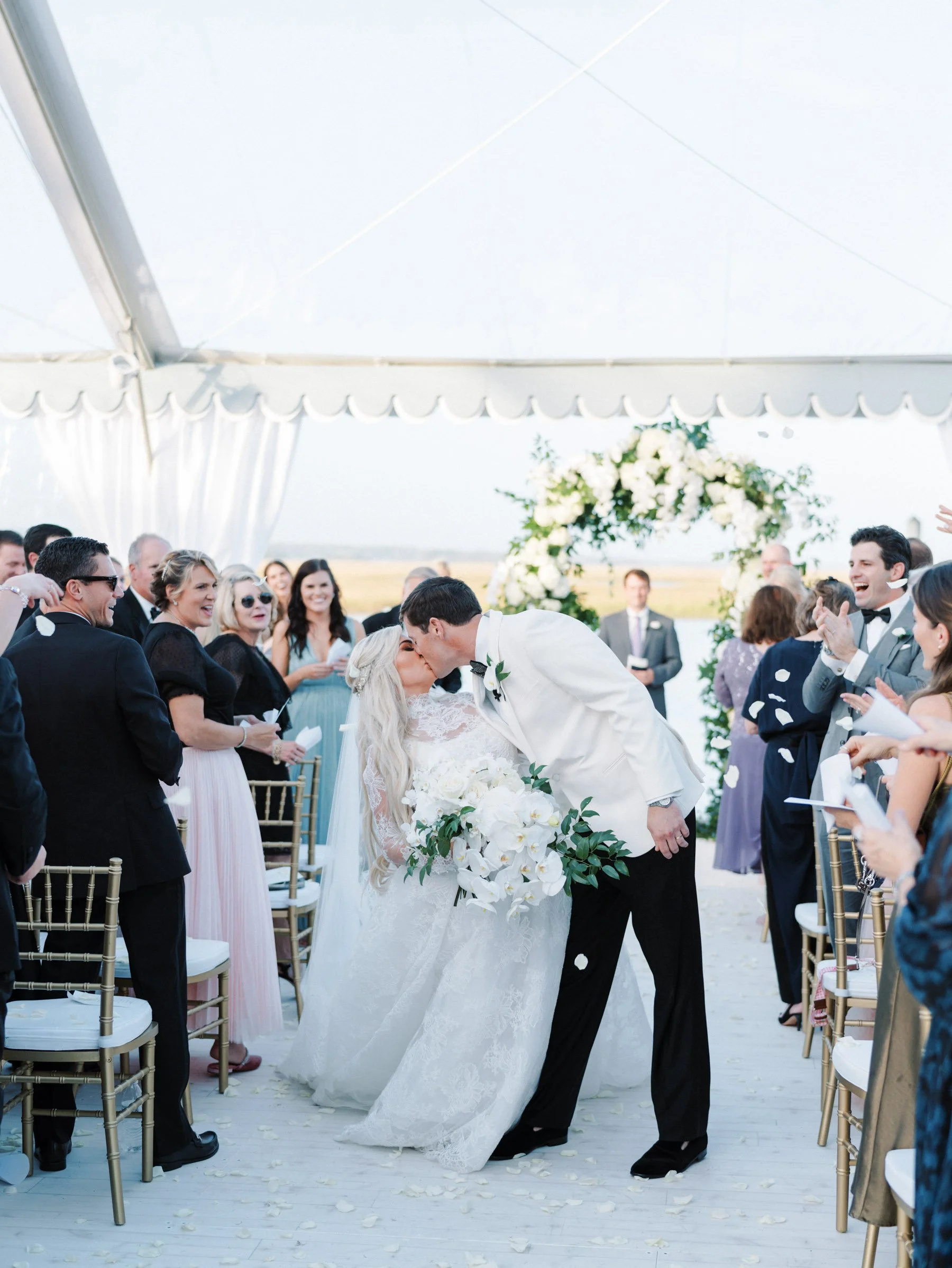 A bride and groom kissing during their wedding ceremony under a white tent, surrounded by guests clapping and throwing white flower petals. The bride is holding a large bouquet of white flowers, and there is a floral arch behind them.