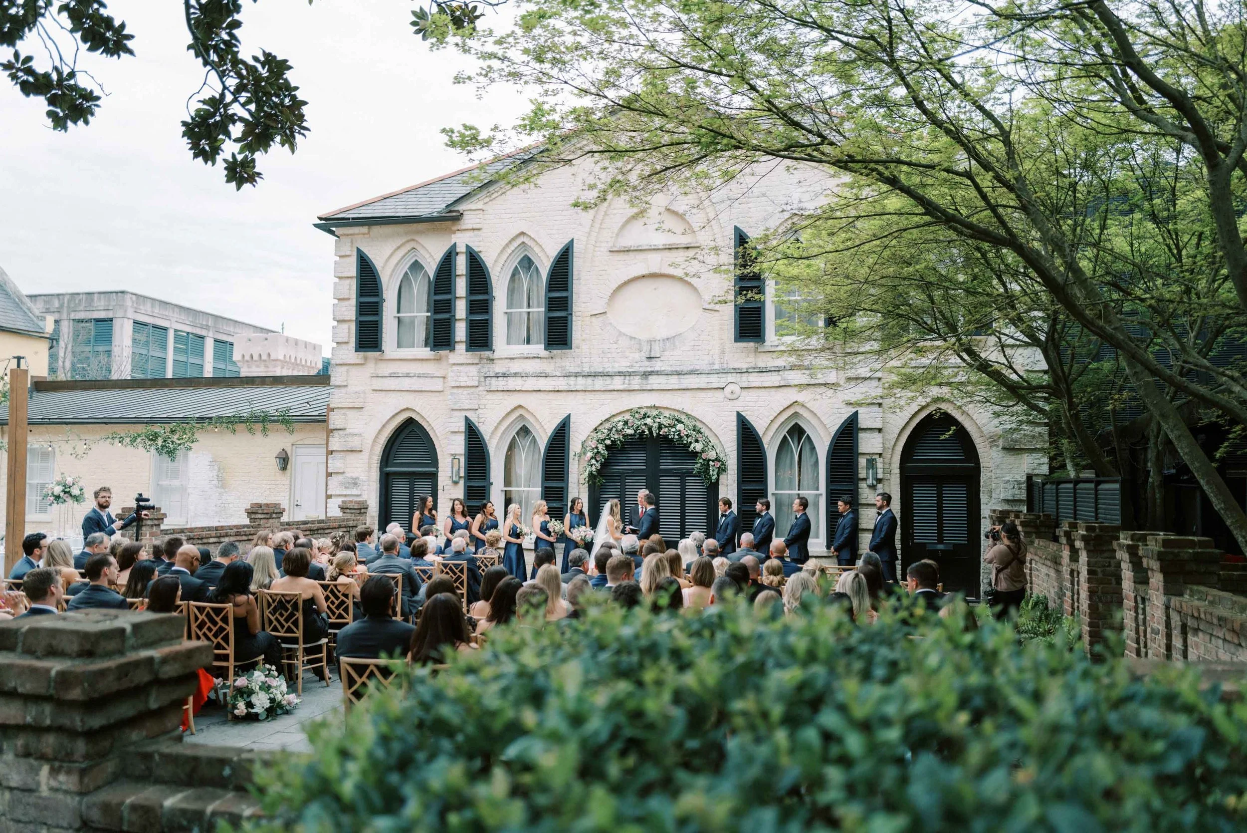 Outdoor wedding ceremony taking place in front of a historic white brick building with arched windows and black shutters. The bride and groom are standing under a floral arch, surrounded by bridesmaids and groomsmen, with seated guests watching. A cameraman is filming, and a photographer is taking pictures on the right.