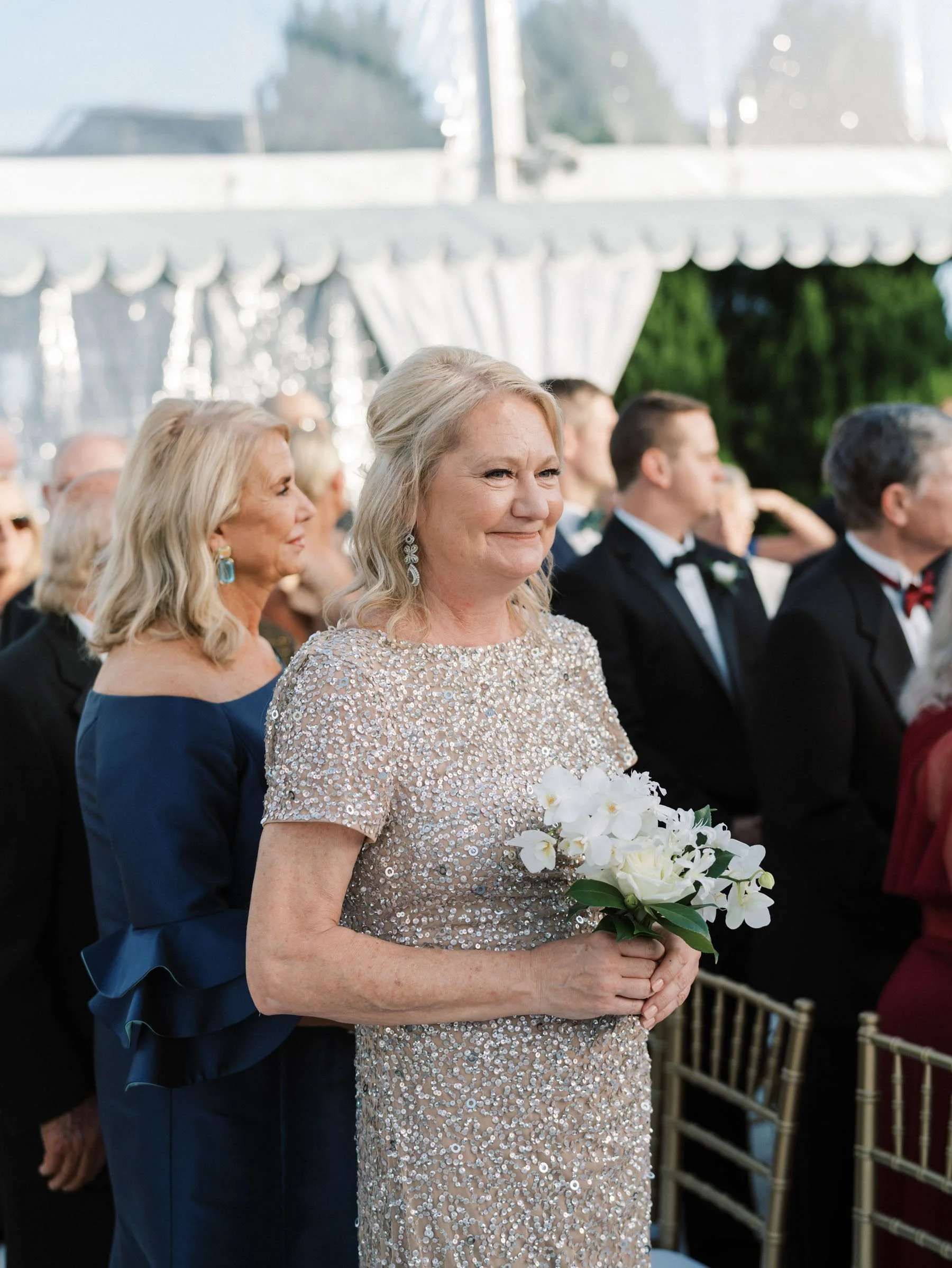 Woman in a sparkling beige gown holding a white flower bouquet at a formal event, possibly a wedding, with other elegantly dressed guests in the background under a white tent canopy.