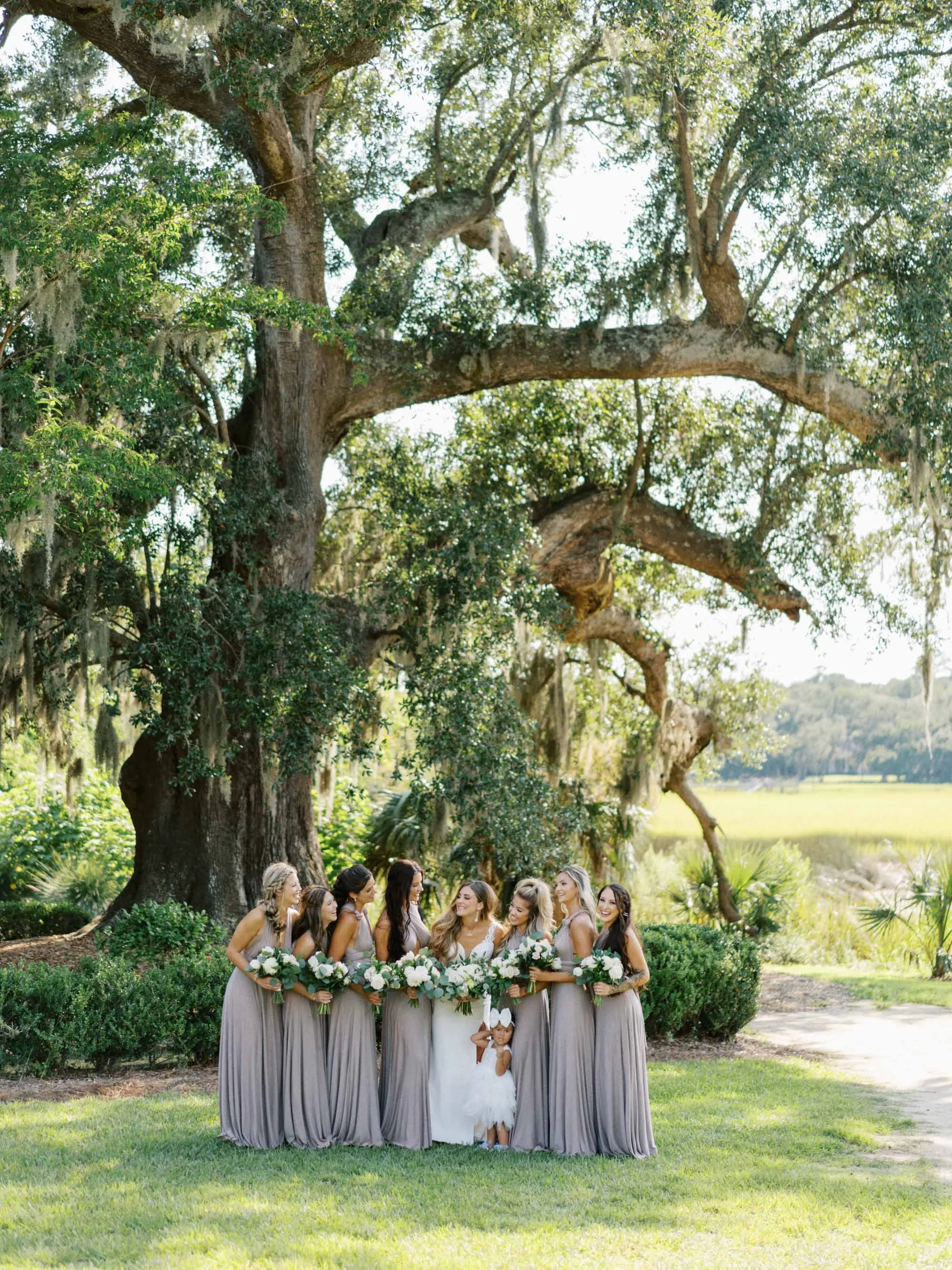 A wedding party posed outdoors under a large tree with lush greenery, including nine women, a young girl in a white dress, all holding bouquets, with the bride in the center at Boone Hall in Charleston.