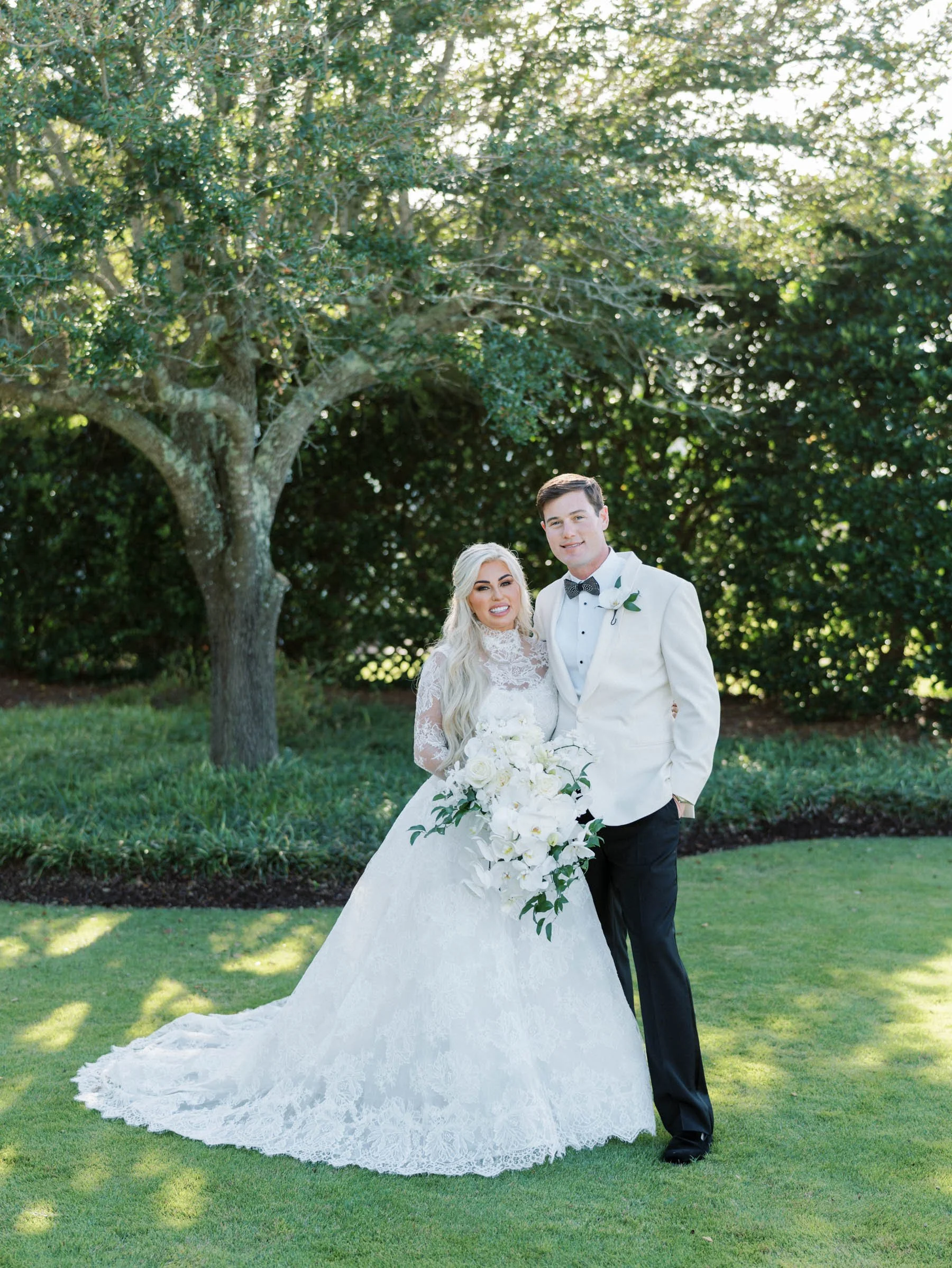 A bride and groom posing outdoors on their wedding day under a large tree, with green grass and shrubbery in the background. The bride is wearing a white lace wedding dress and holding a bouquet of white flowers, while the groom is dressed in a white