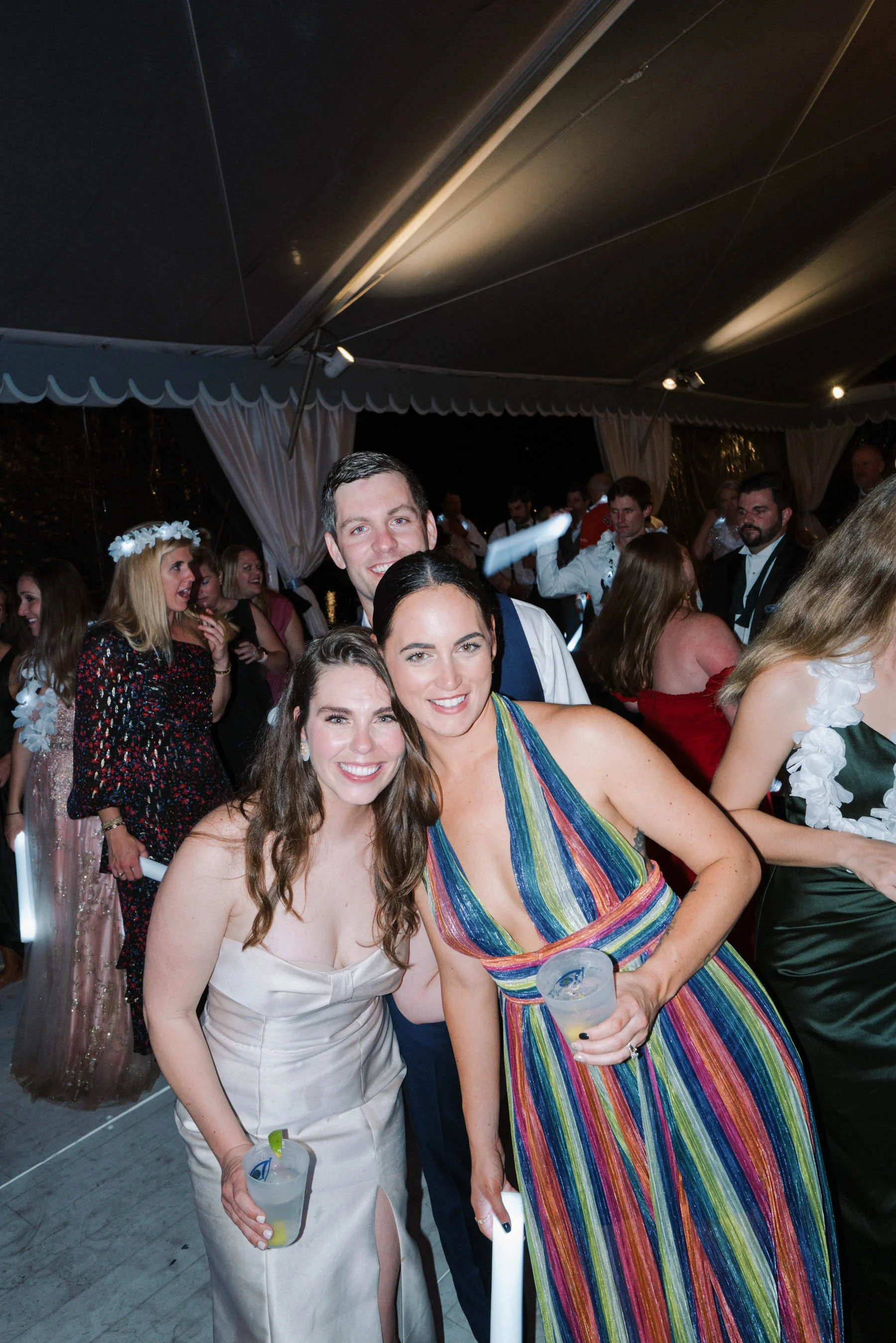 People dancing and celebrating at a social event or party inside a tent with string lights, with some holding drinks, smiling, and dressed in formal or semi-formal attire.