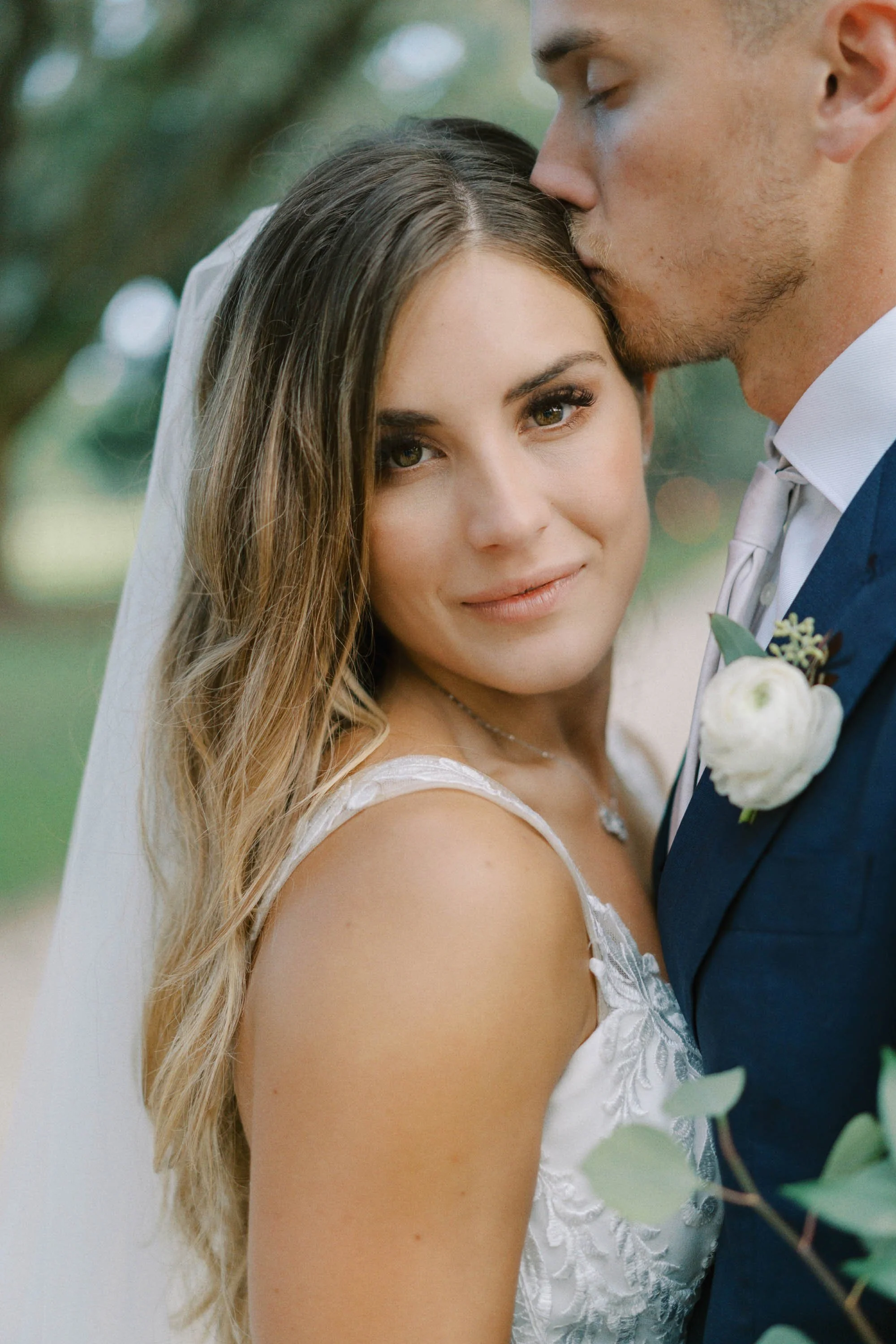 A bride and groom on their wedding day, with the groom kissing the bride on her forehead. The bride has long, wavy brown hair and is wearing a white lace wedding dress. The groom is dressed in a navy suit with a white shirt, gray tie, and a white boutonniere, with a blurred green outdoor background.