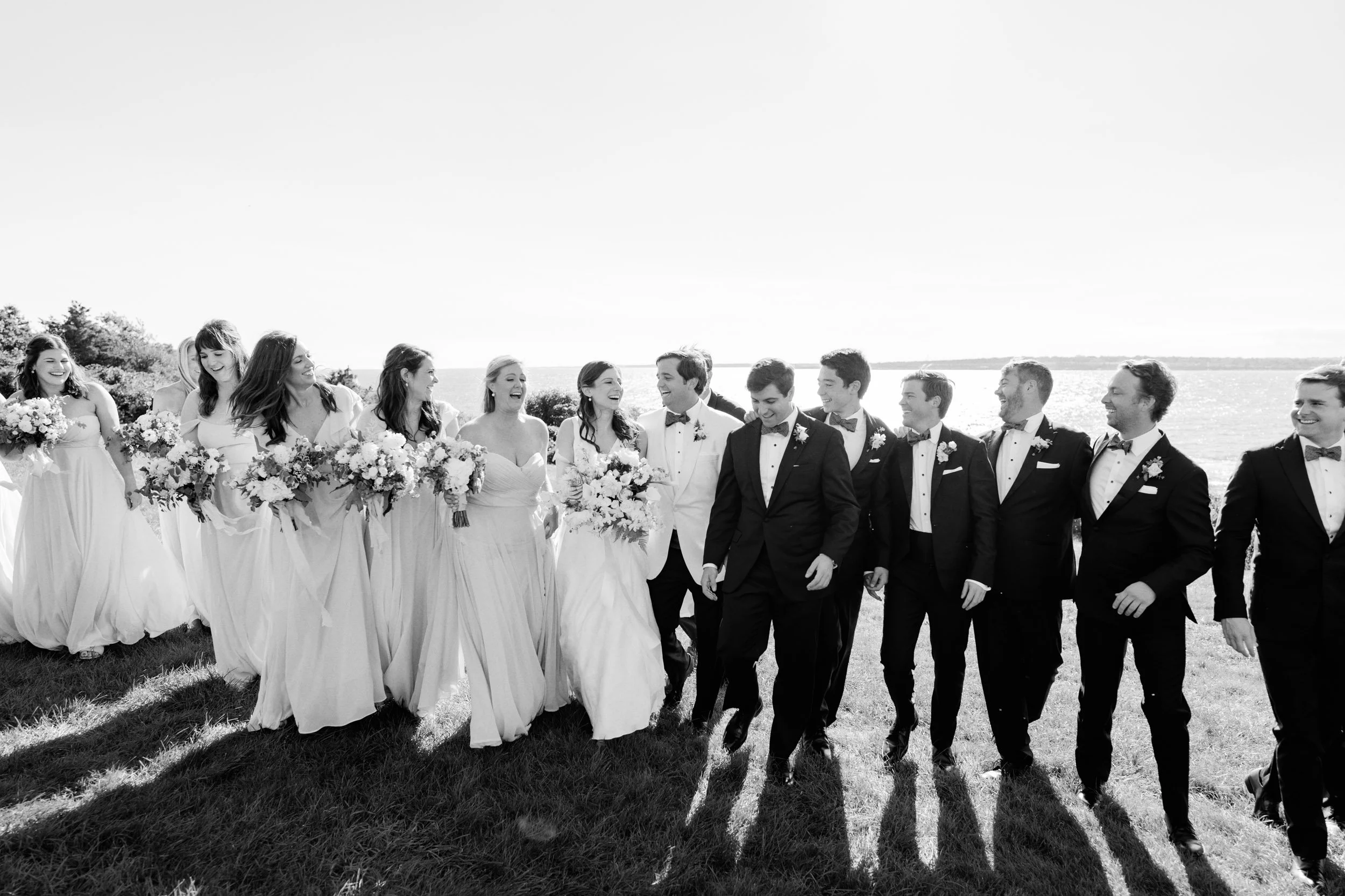 Wedding party walking outdoors near water, women in dresses holding bouquets, men in tuxedos