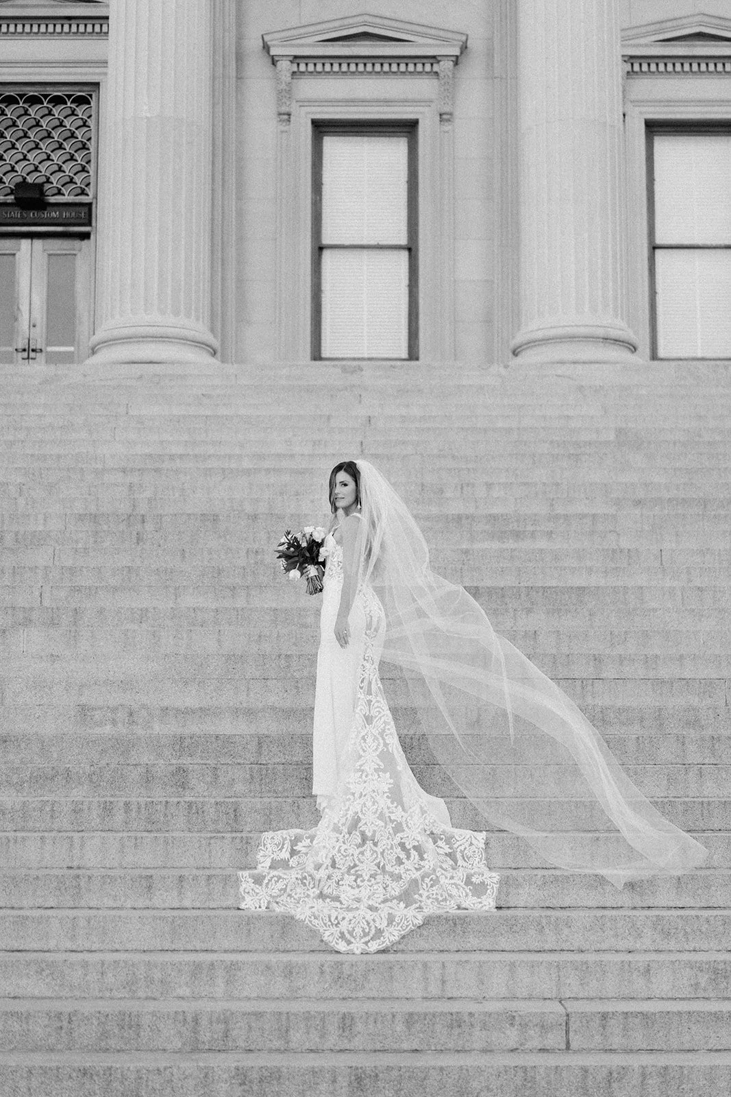 Black-and-white photo of a bride standing on steps in front of a neoclassical building with large columns, holding a bouquet of flowers, wearing a wedding dress with lace details and a veil.