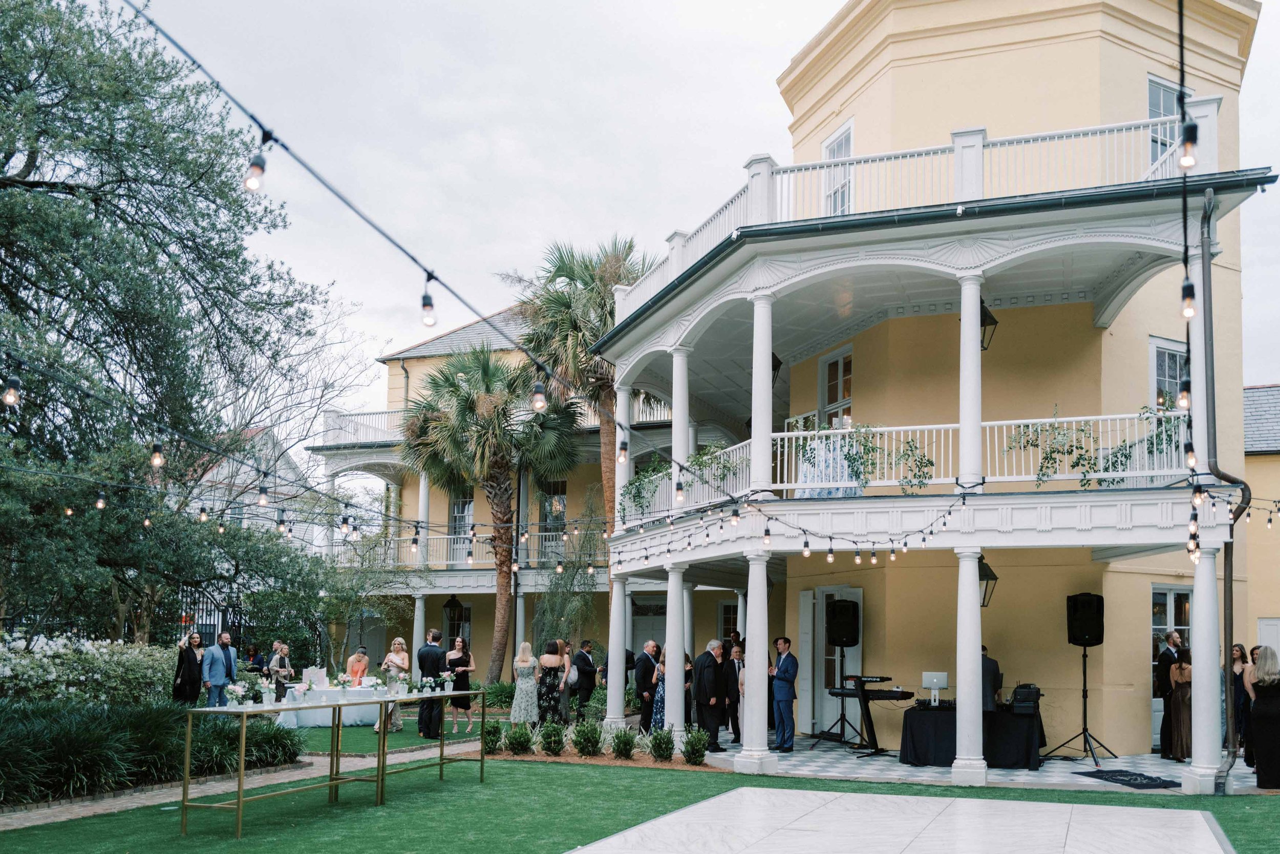 Guests mingle at an outdoor event on a lawn in front of a yellow multi-story house with white balconies and columns. String lights are hanging overhead, and there is a table with refreshments on the left.