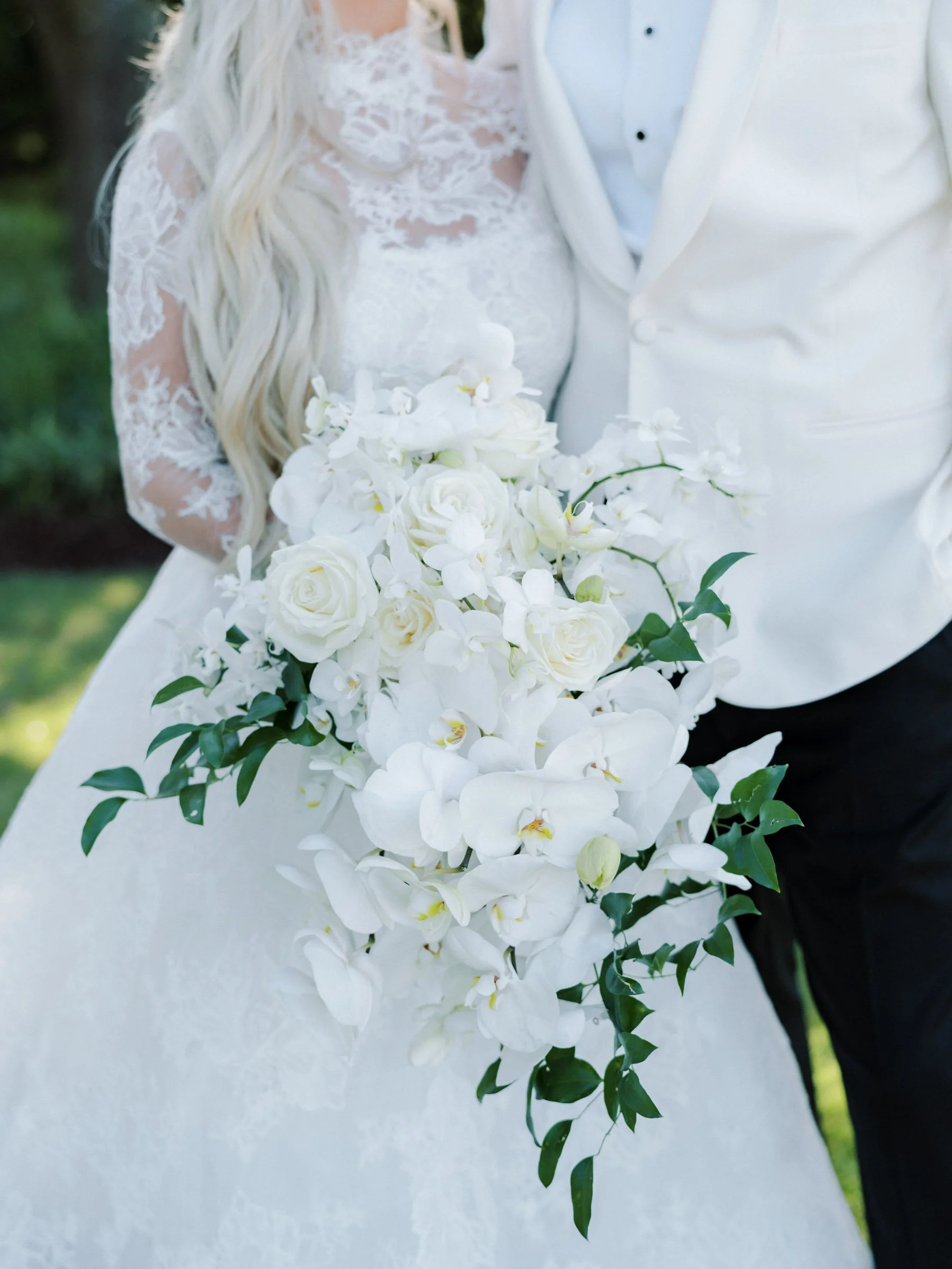 Bride in white wedding dress holding a cascading bouquet of white roses, orchids, and greenery, standing next to groom in white tuxedo jacket and black pants.