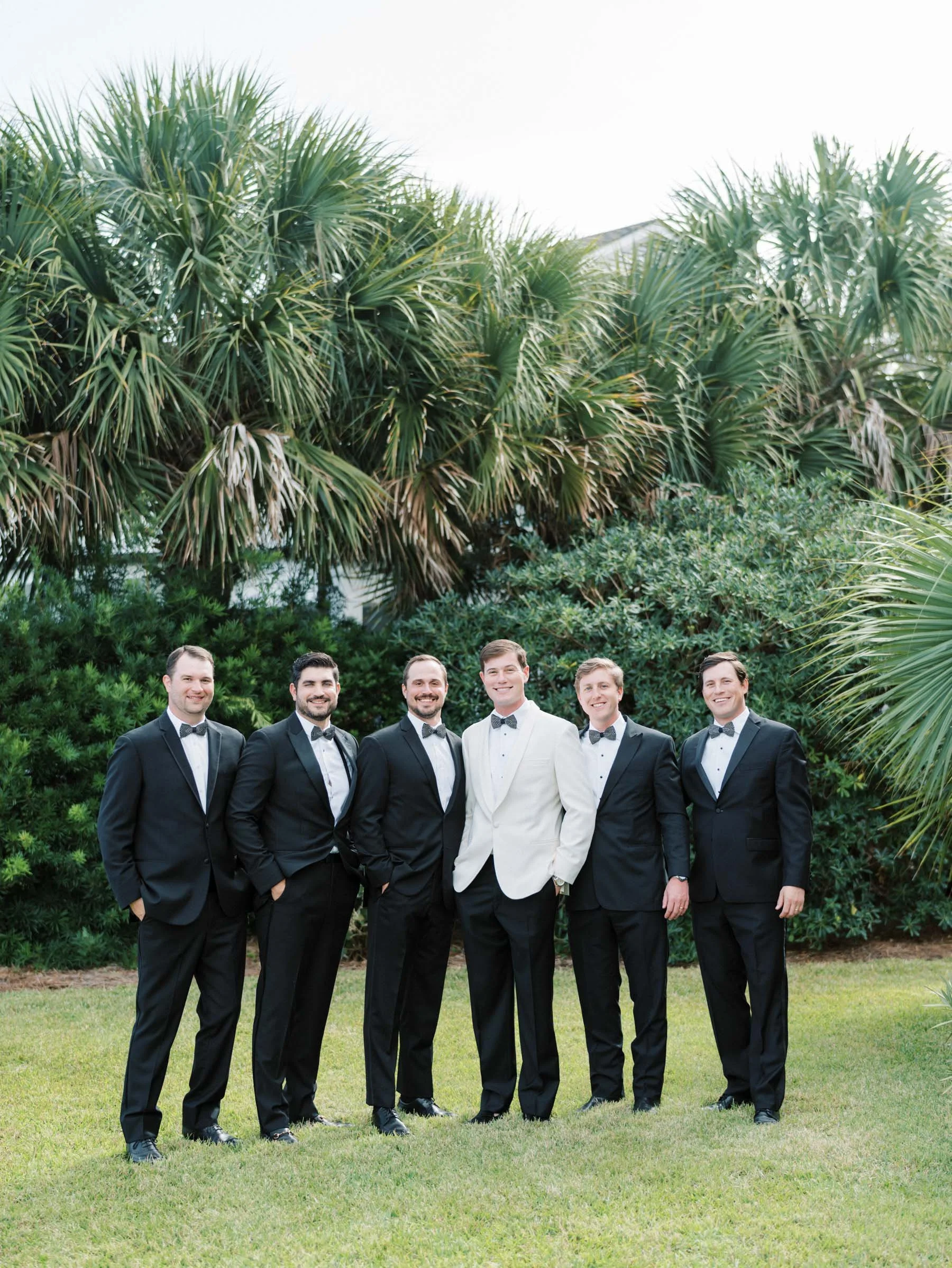 Group of six men in suits, with one in a white tuxedo jacket, standing outside on grass with large palm trees and bushes in the background.