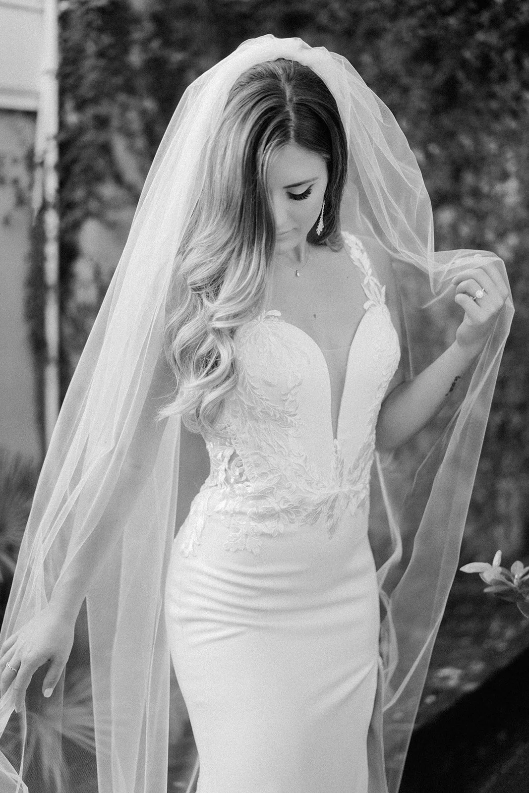 A bride in a wedding dress and veil outdoors, looking down and holding part of her veil.