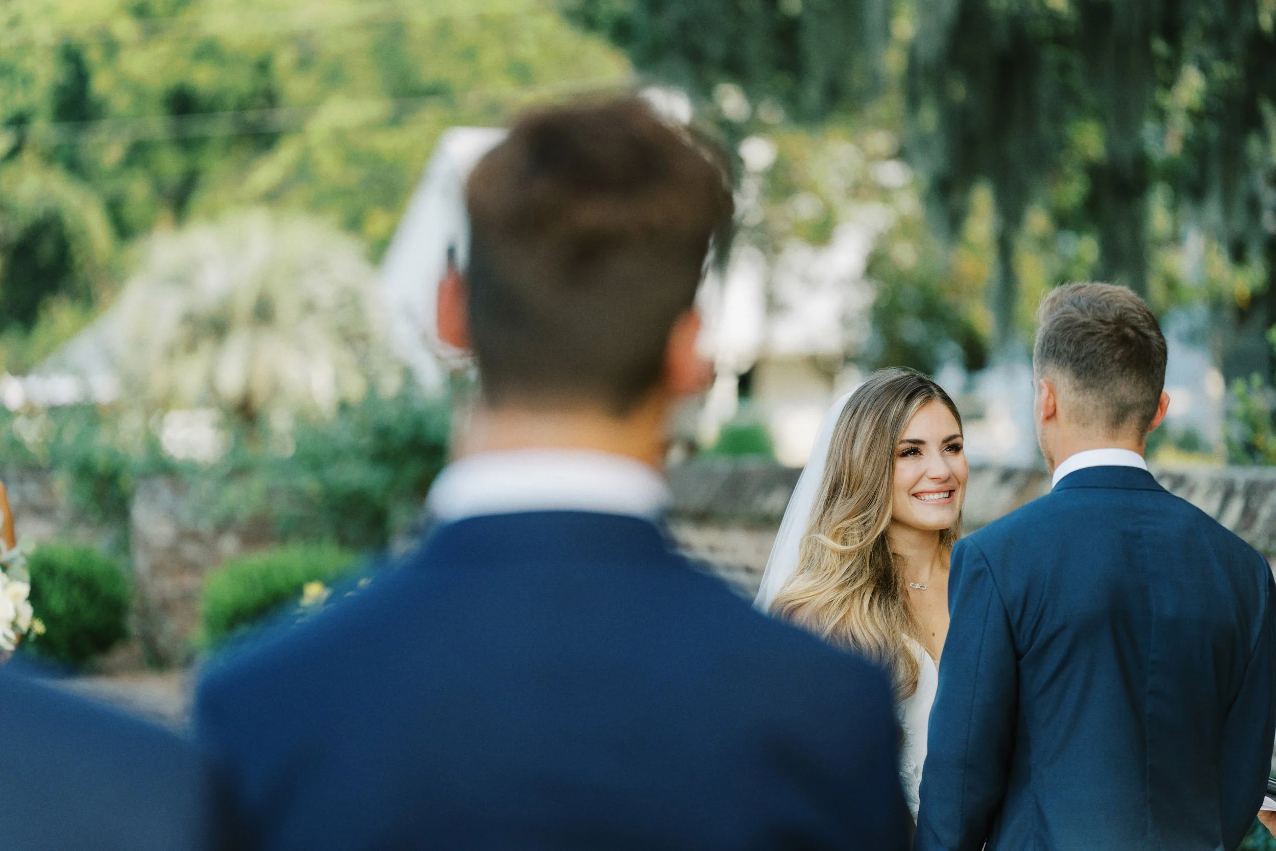 A bride smiling at a groom during an outdoor wedding ceremony with trees and greenery in the background