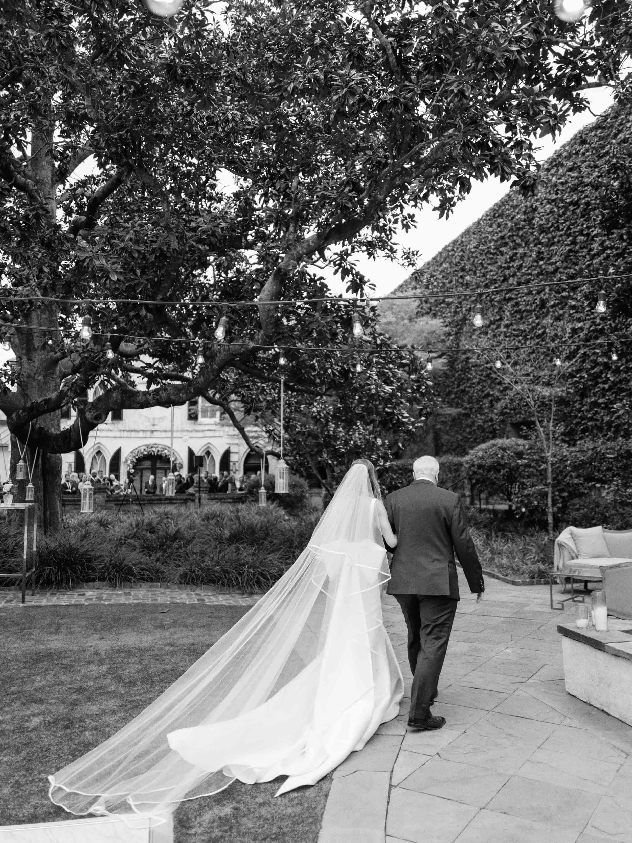 A bride and an older man, possibly her father, walking in an outdoor wedding setting. The bride is wearing a long veil and wedding gown, and the scene is decorated with string lights, trees, and a hedge in the background.