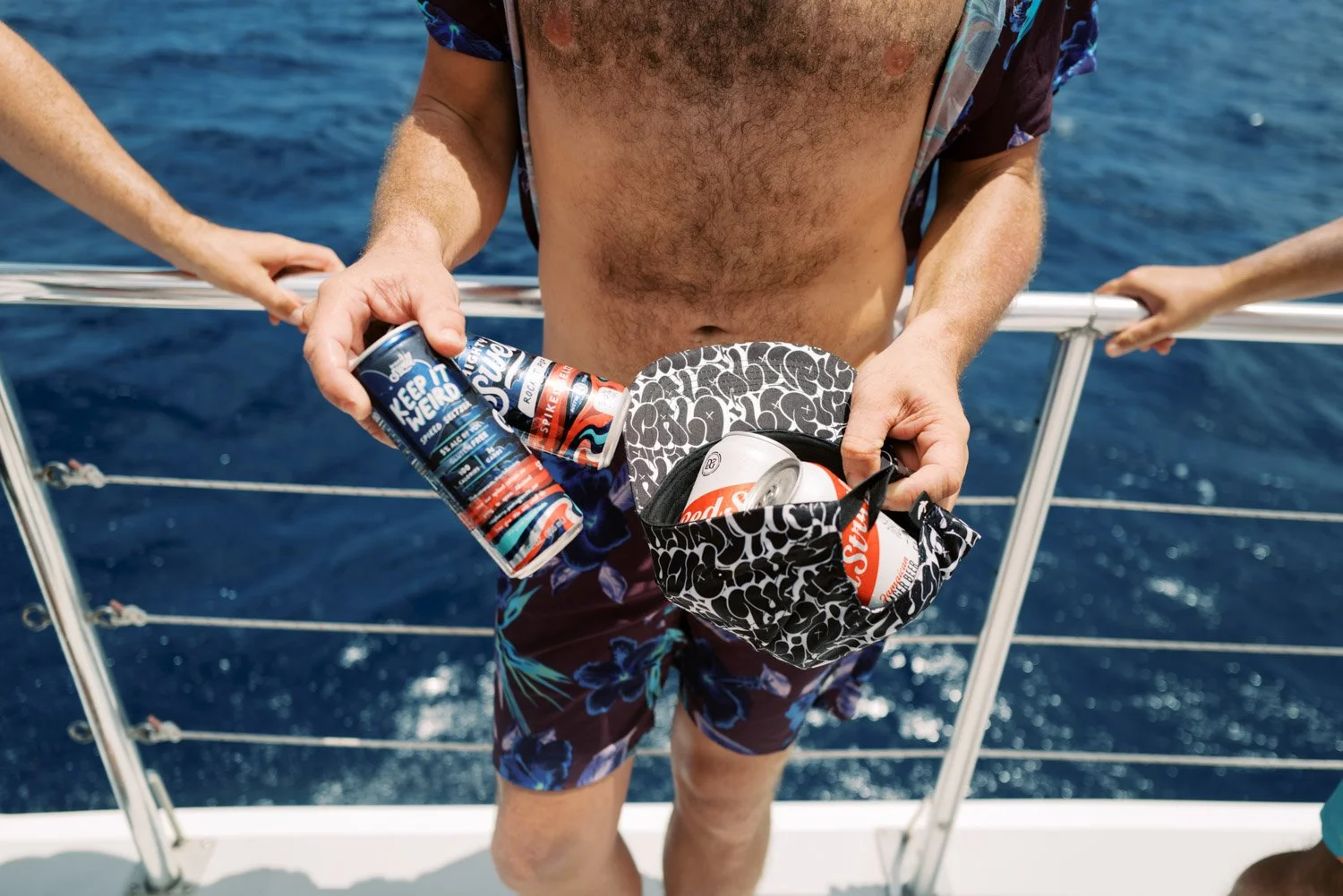 A man on a boat holding a can of beer and placing other cans in a black and white patterned small bag, with the ocean in the background.