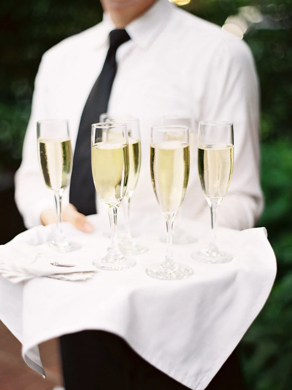 A waiter holding a white tray with six glasses of champagne, dressed in a white shirt and black tie, with a blurred greenery background.