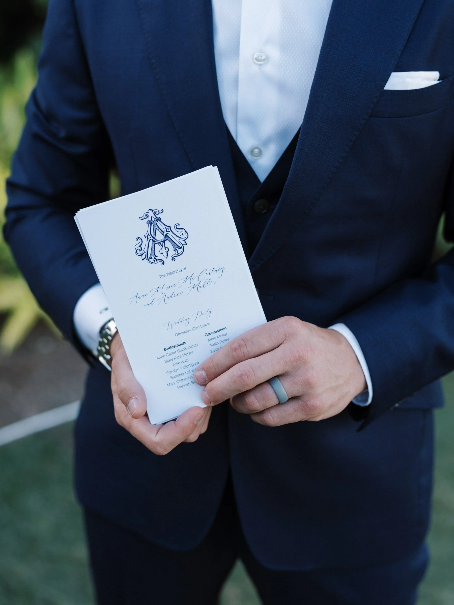 A man in a navy blue suit holding a wedding program with a monogram at the top, standing outdoors.