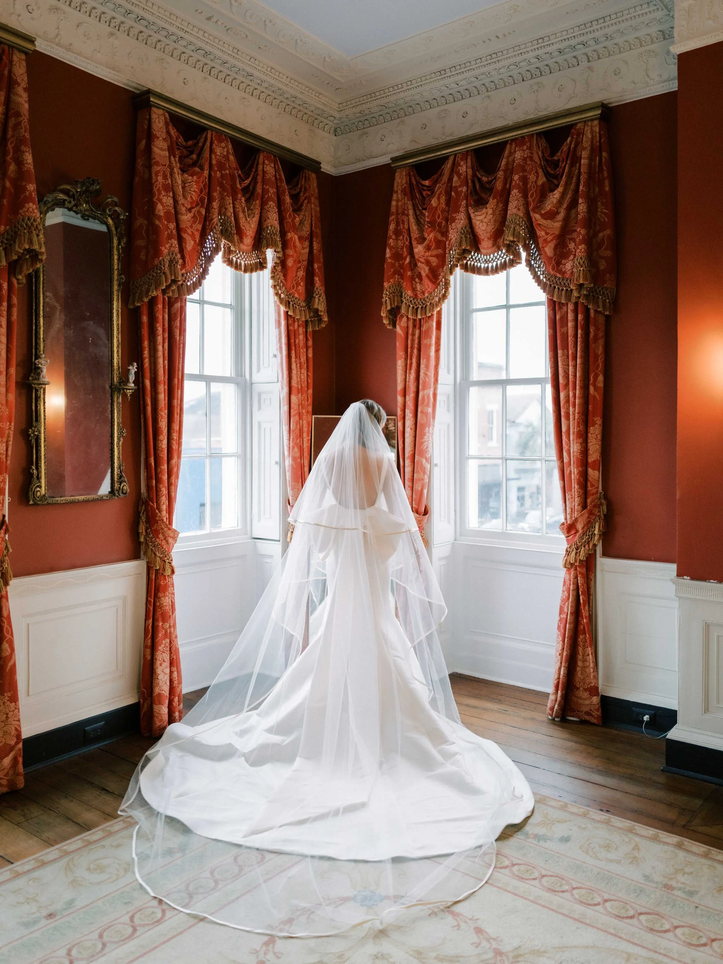 A bride in a white wedding dress with a long train and veil standing in an elegant room with red walls, ornate curtains, large windows, and a vintage mirror.