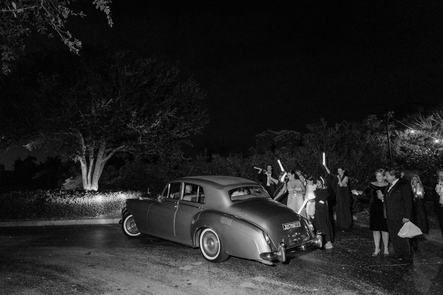 Vintage black and white photo of a wedding celebration at night with a classic car and a group of people holding glowing sticks or sparklers.