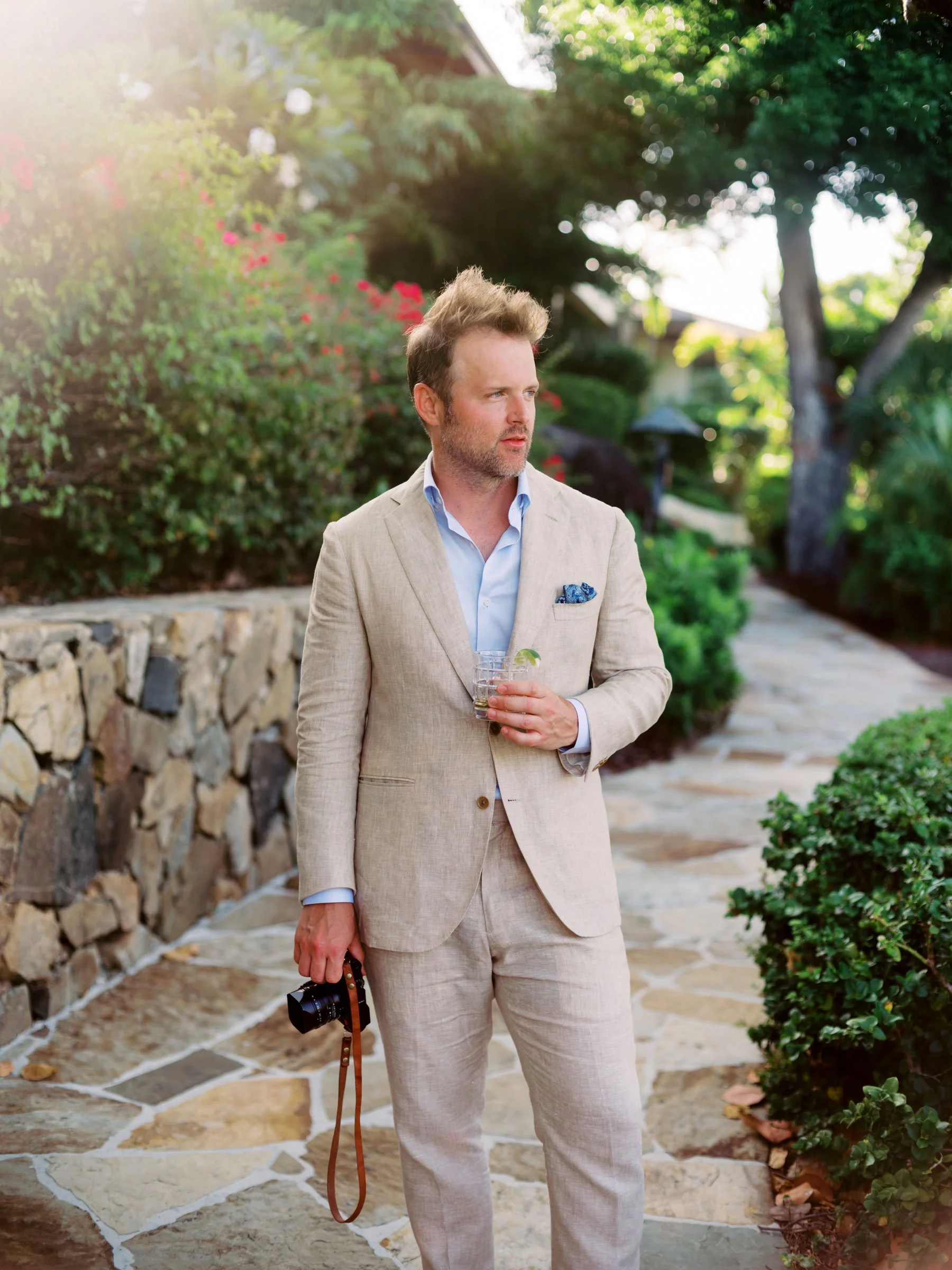 A man in a beige suit holding a camera in one hand and a glass of drink in the other, standing on a stone pathway in a lush garden.