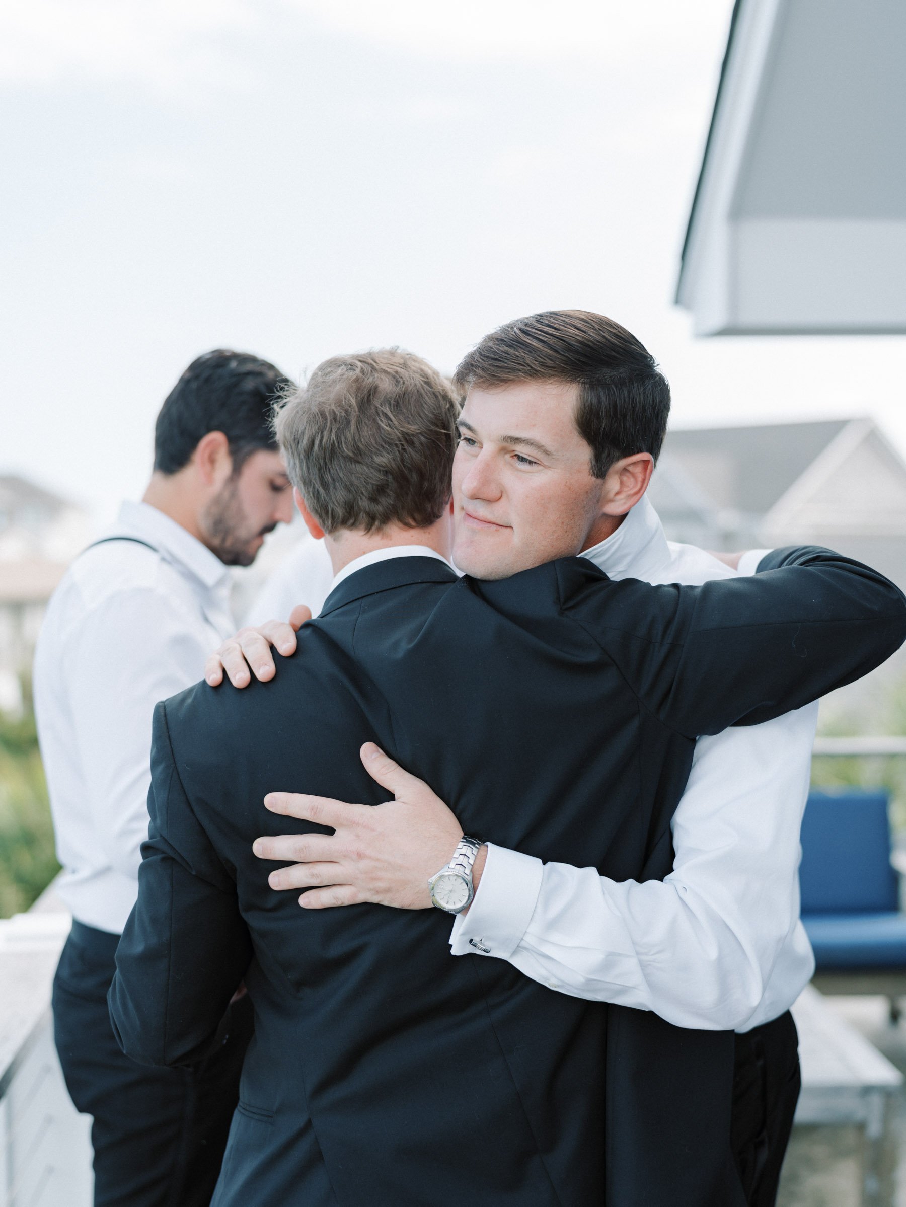 Two men in suits hugging, one with a watch on his left wrist, outdoors in a modern setting.
