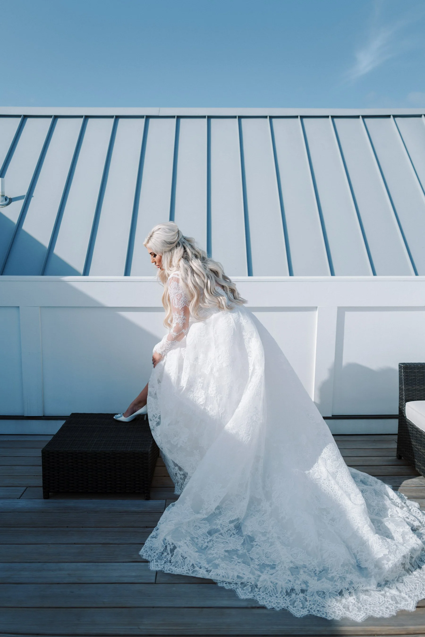 Bride in a white lace wedding gown sitting on a wicker ottoman on a wooden deck, adjusting her shoe, with a white building and a clear blue sky in the background.