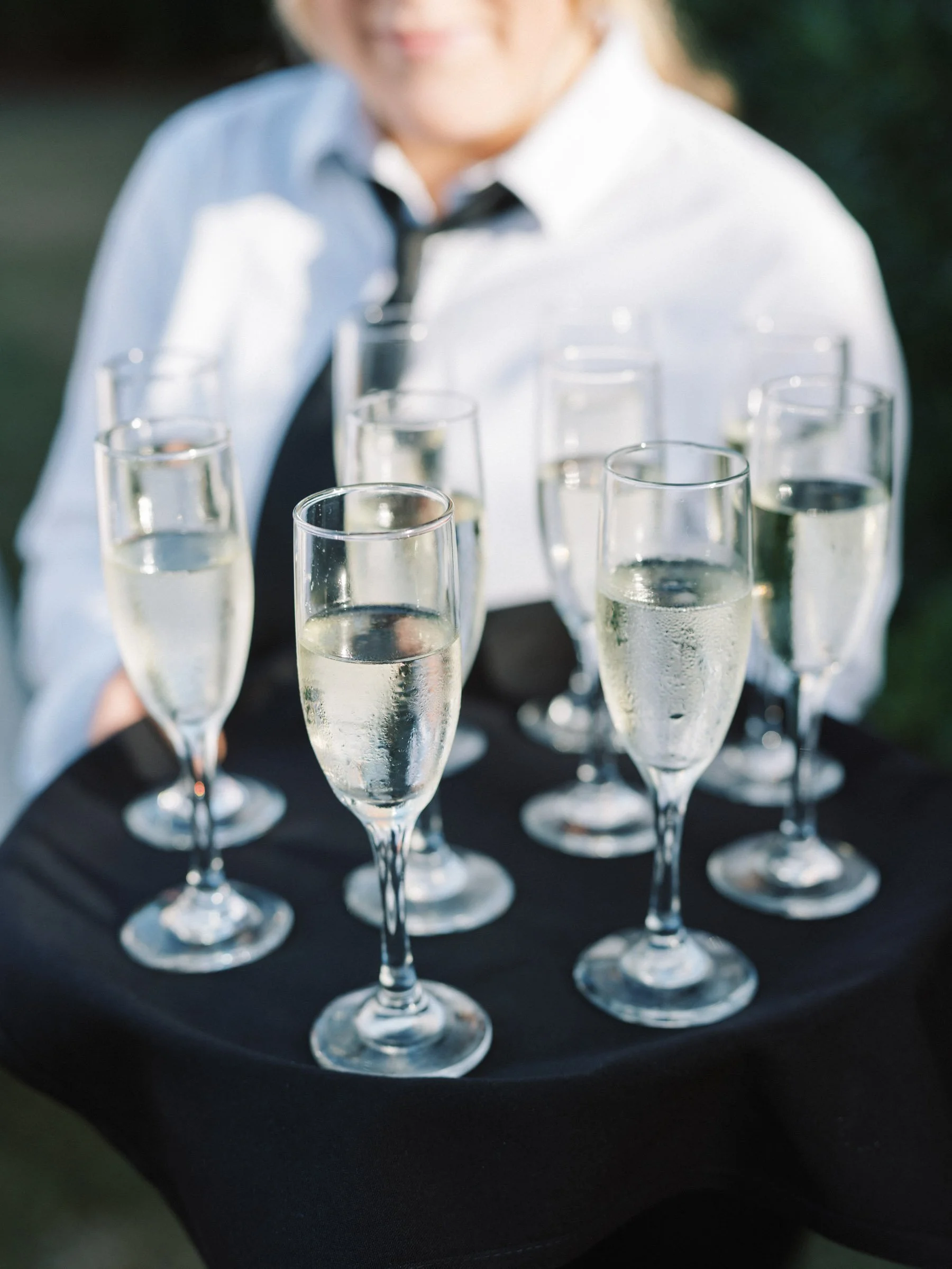 A person dressed in a white shirt and black tie holding a tray with eight glasses of champagne.