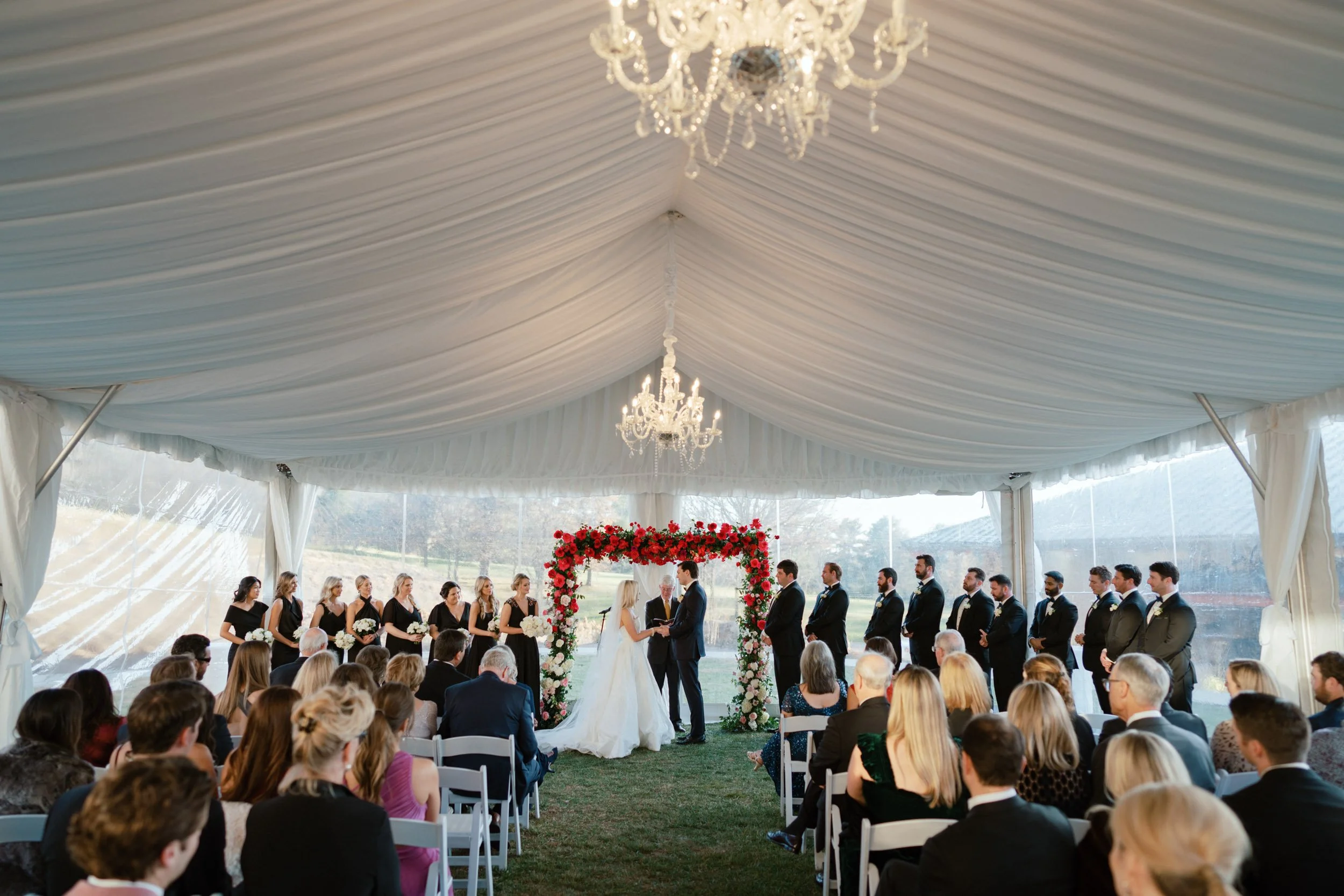 A wedding ceremony taking place under a large white tent with a floral arch at the altar. The bride and groom are standing facing each other, holding hands. Bridesmaids in black dresses and groomsmen in black suits are lined up on either side of the 