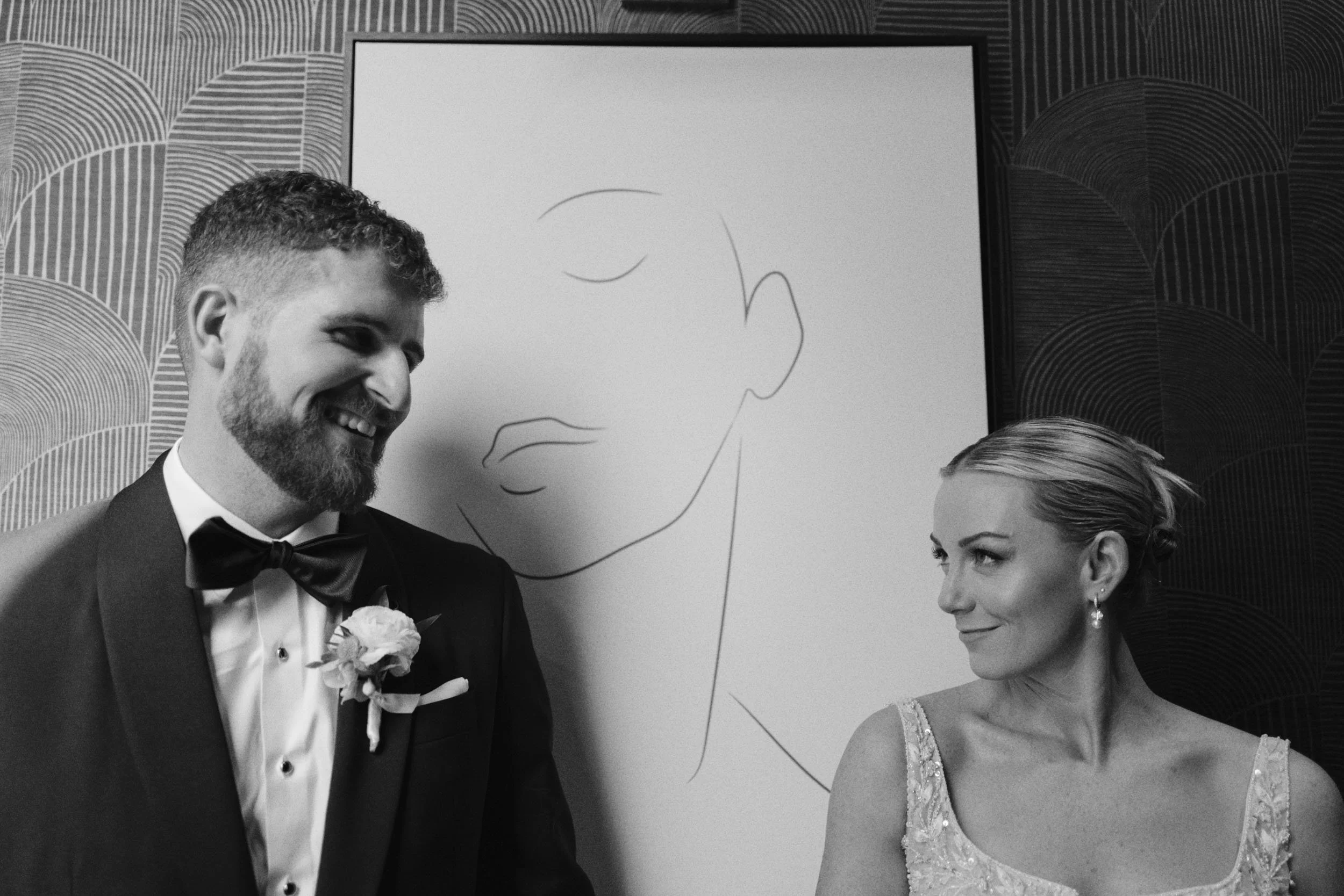 A black and white photo of a bride and groom smiling at each other, standing in front of a minimalist line art portrait on a whiteboard.