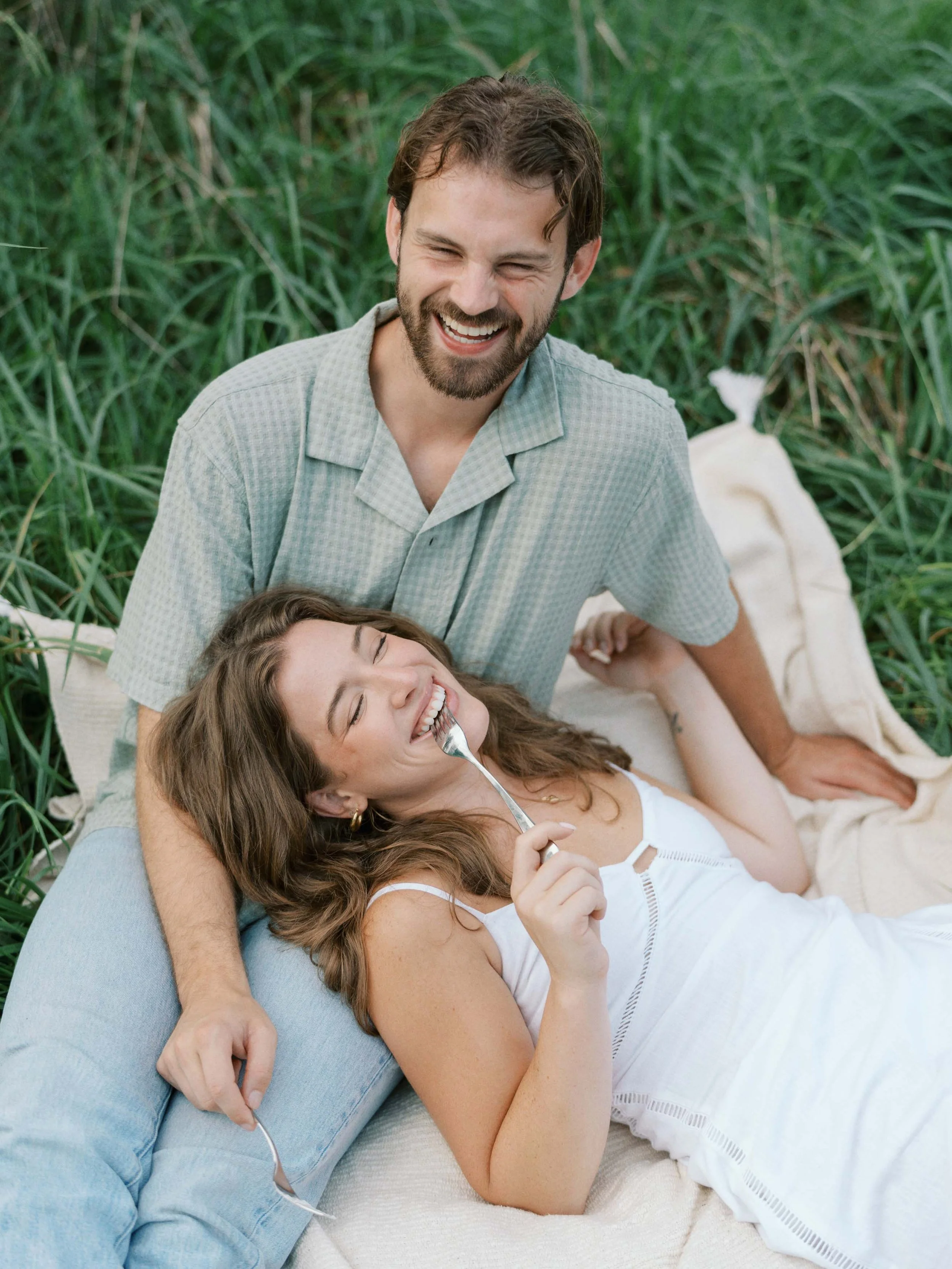 A young couple, a man and a woman, enjoying a picnic in a grassy outdoor area, laughing and smiling.