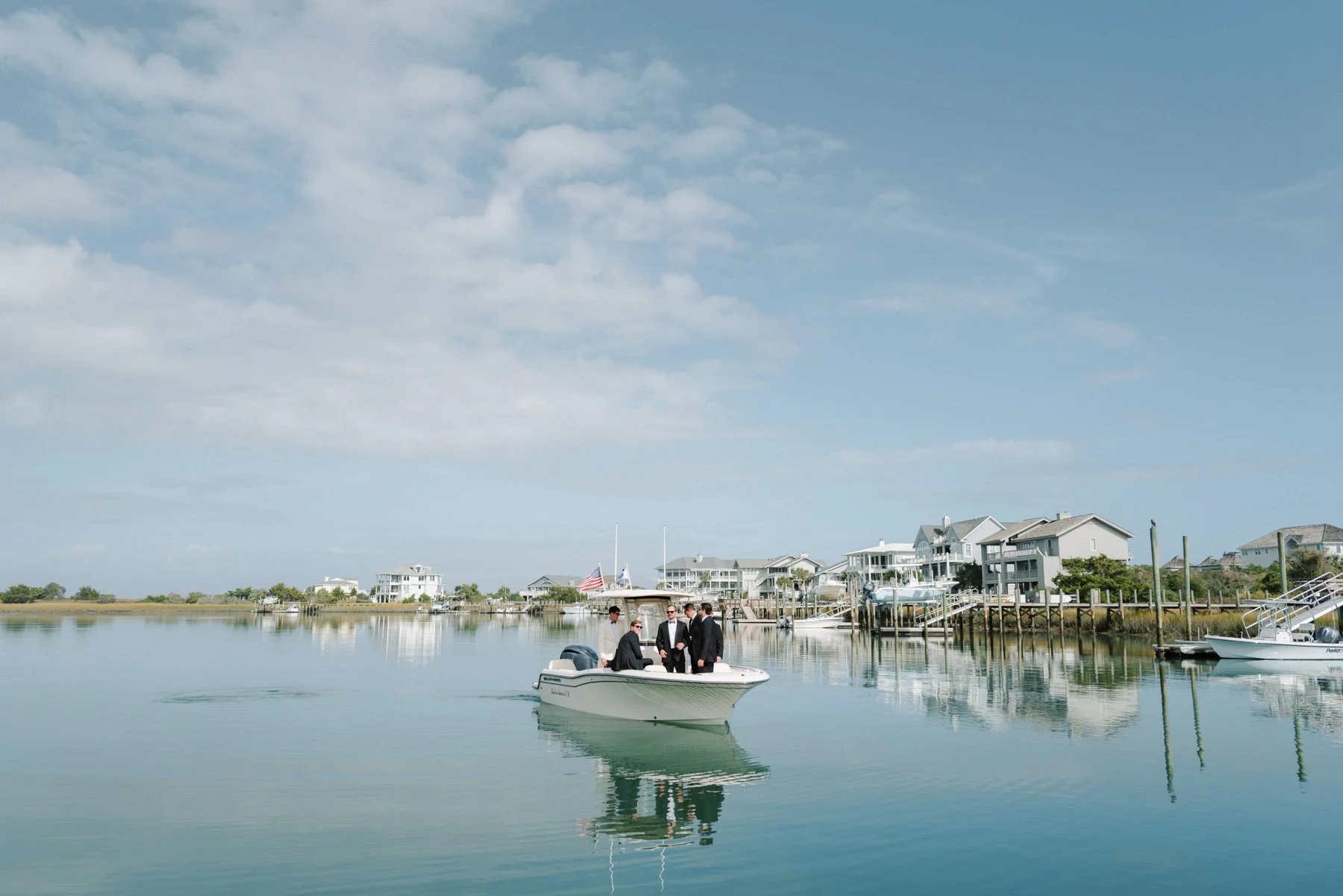 A boat with four men in suits on calm water near houses on the shoreline.
