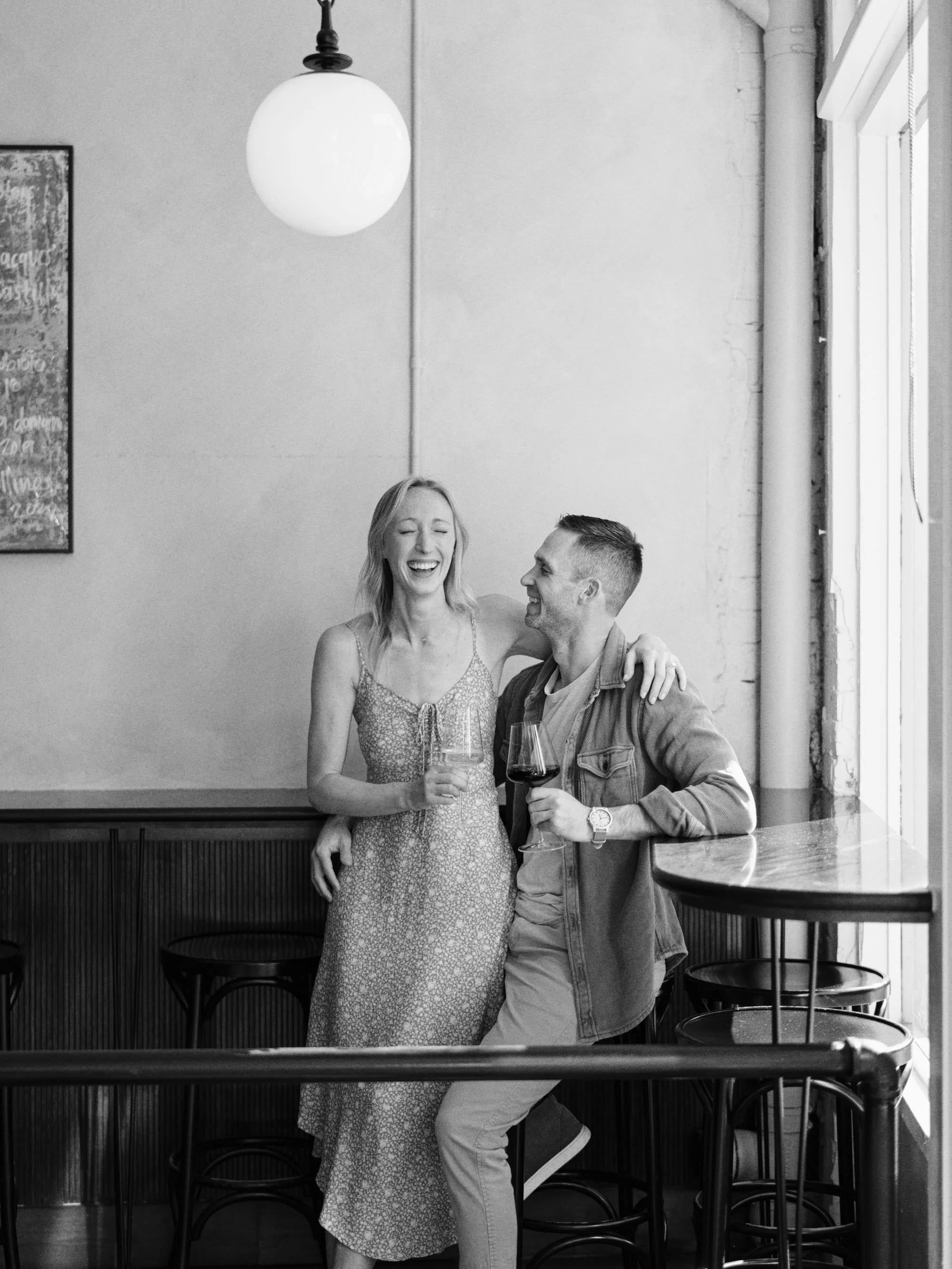 A black and white photo of a woman and a man with glasses of wine, smiling and sharing a joyful moment in a cafe or bar.