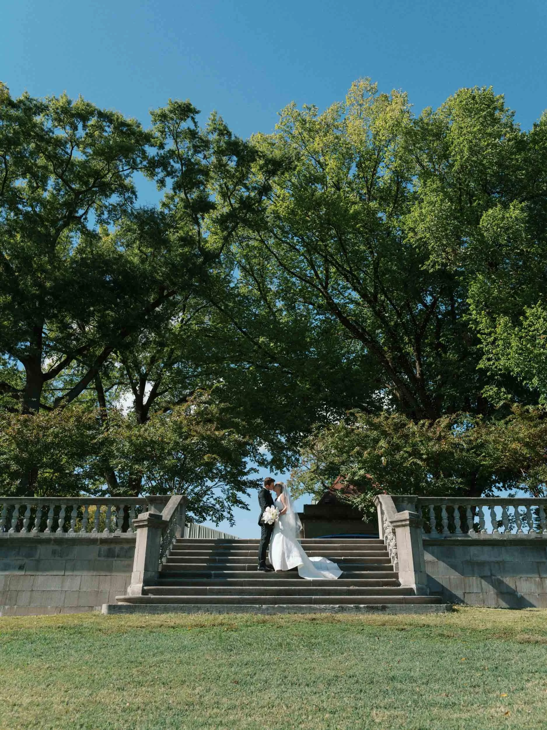 A bride and groom in wedding attire standing on outdoor stairs under a large green tree, with a clear blue sky in the background.