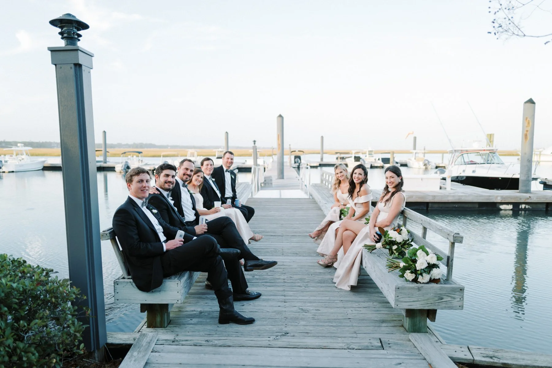 A wedding party sitting on a wooden dock by a marina with boats in the background. Men in tuxedos and women in white dresses are smiling and looking at the camera. Bridal bouquets are placed on the benches.