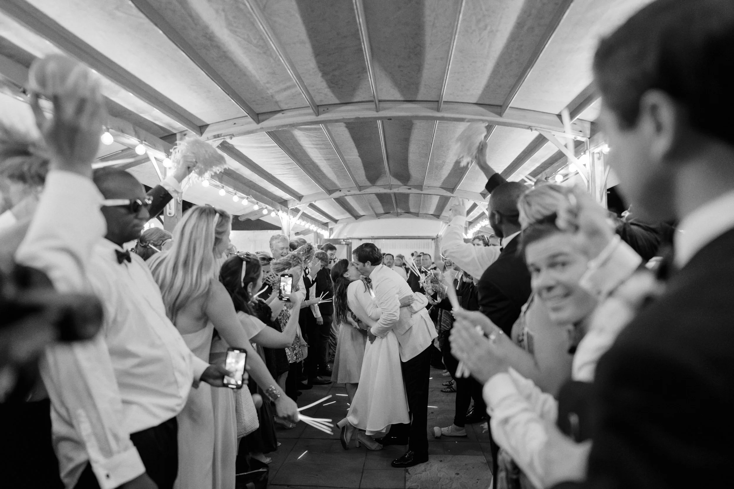 Black and white photo of a wedding reception with a bride and groom kissing in the center, surrounded by guests holding sparklers, some taking photos, in a decorated outdoor pavilion with string lights.