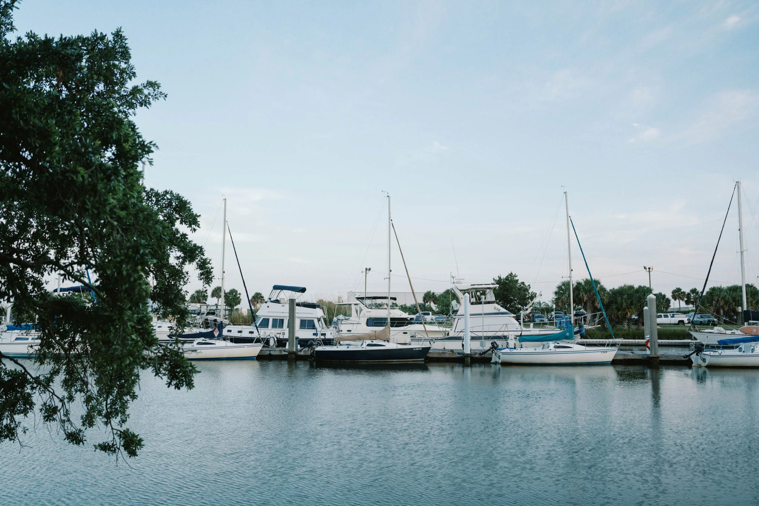 Marina with several sailboats and motorboats docked along the pier, with water in the foreground and a partly cloudy sky above.