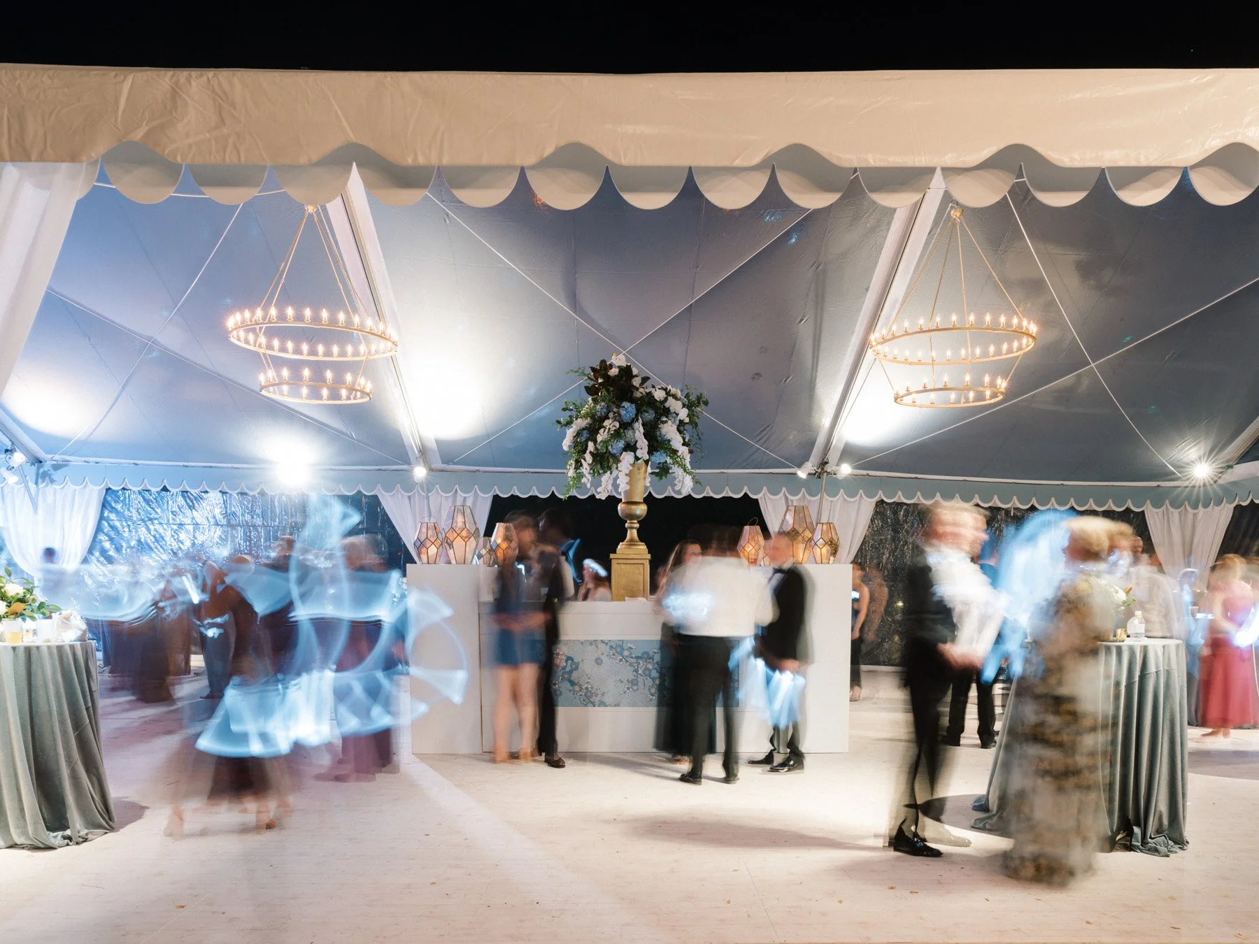 Elegant wedding reception under a large white tent with chandeliers, floral centerpiece, and guests mingling.