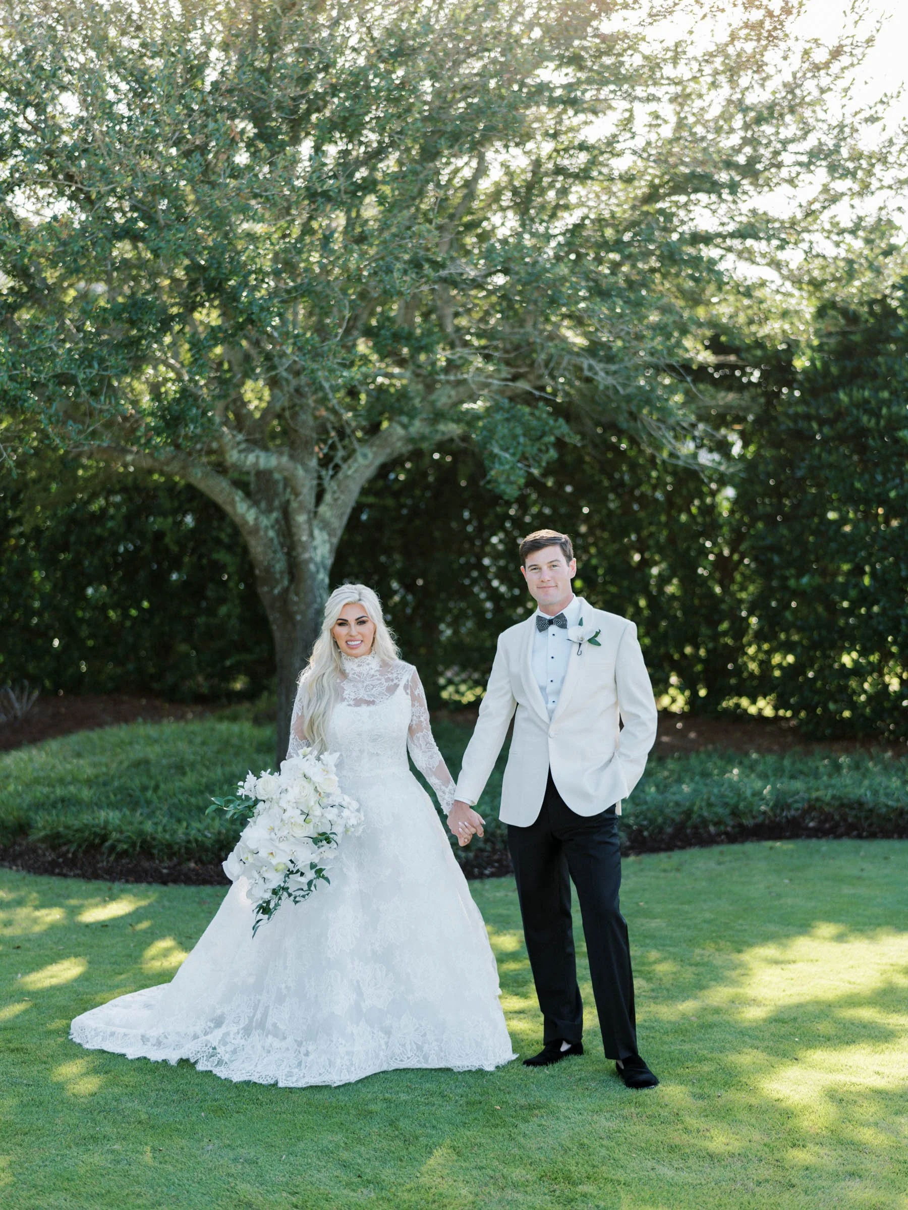 Bride and groom holding hands outdoors on wedding day, standing on grass with a tree and greenery in the background.
